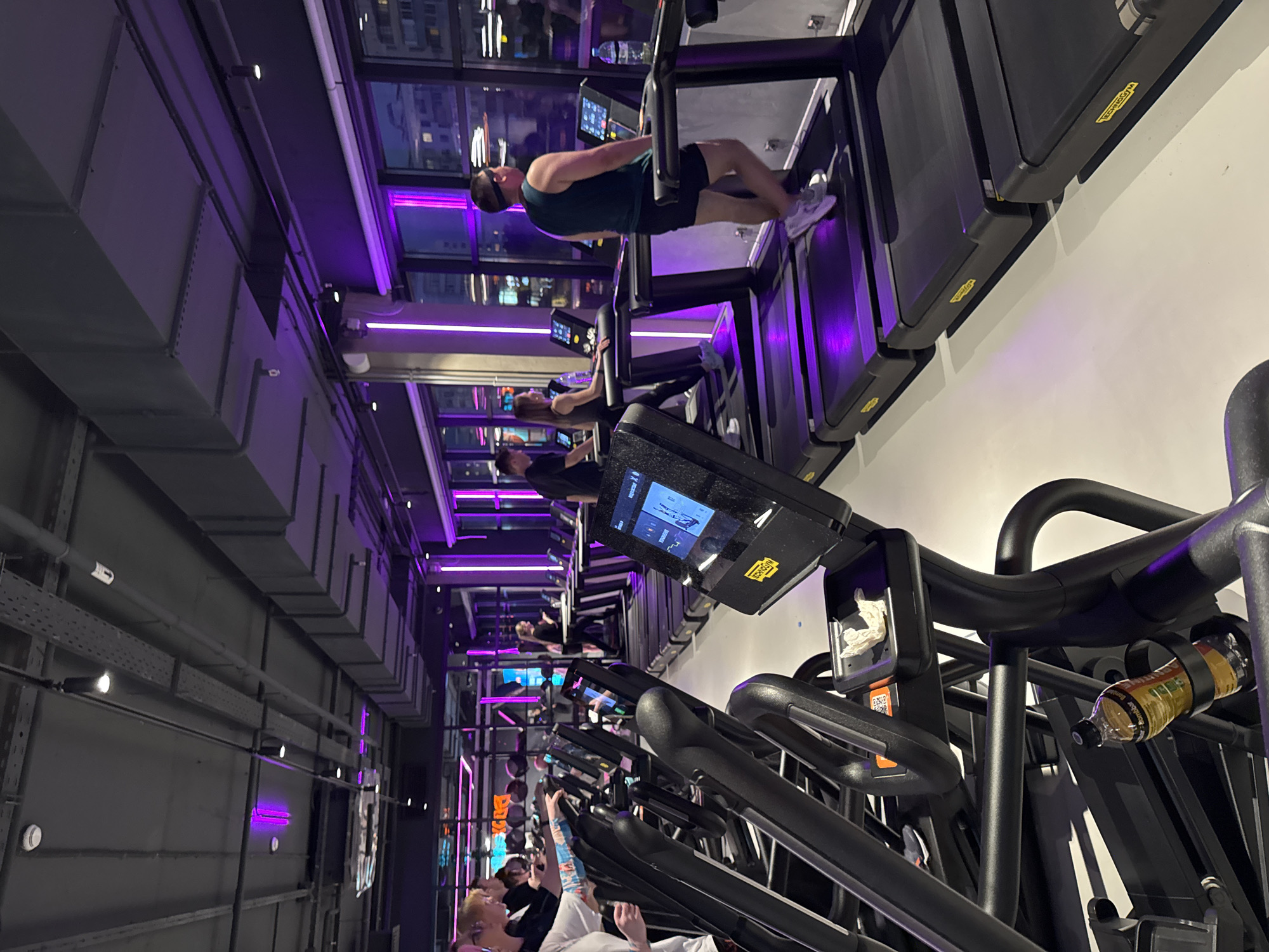 A gym in Moscow, Russia is seen with a row of people using treadmills. The treadmills are black and have digital screens. The people are dressed in athletic wear. The gym has a modern design with purple lighting. The ceiling has exposed pipes and ducts. Outside the windows are other buildings and city lights.