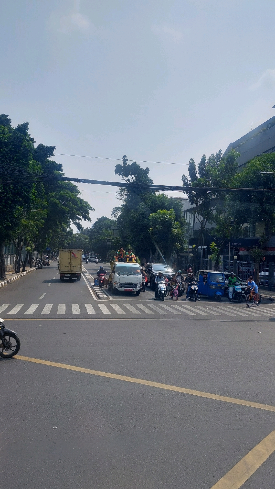 The image captures a daytime street scene at an intersection in East Jakarta, Indonesia. The sky is clear blue with a few wispy clouds. The foreground shows a grey asphalt road with distinct white zebra crossings and a yellow dividing line. A black motorcycle is partially visible on the left side of the frame.
The intersection is populated with various vehicles. A white truck with several individuals in bright yellow shirts standing on its cargo bed is stopped at the intersection. To the right of the truck, a dark-colored SUV and several motorcycles are also present. A blue three-wheeled vehicle, commonly known as a "bajaj" in Indonesia, is seen with a driver and passengers. Further to the right, a person is riding a bicycle. On the opposite side of the road, a yellow box truck is moving away from the camera.
The environment features mature trees lining the sidewalks on both sides of the street. In the background, a modern building with several floors and a glass facade is visible, with "Service" written on its side. Power lines are strung overhead across the width of the street. The overall impression is a busy urban intersection under clear sunny weather.