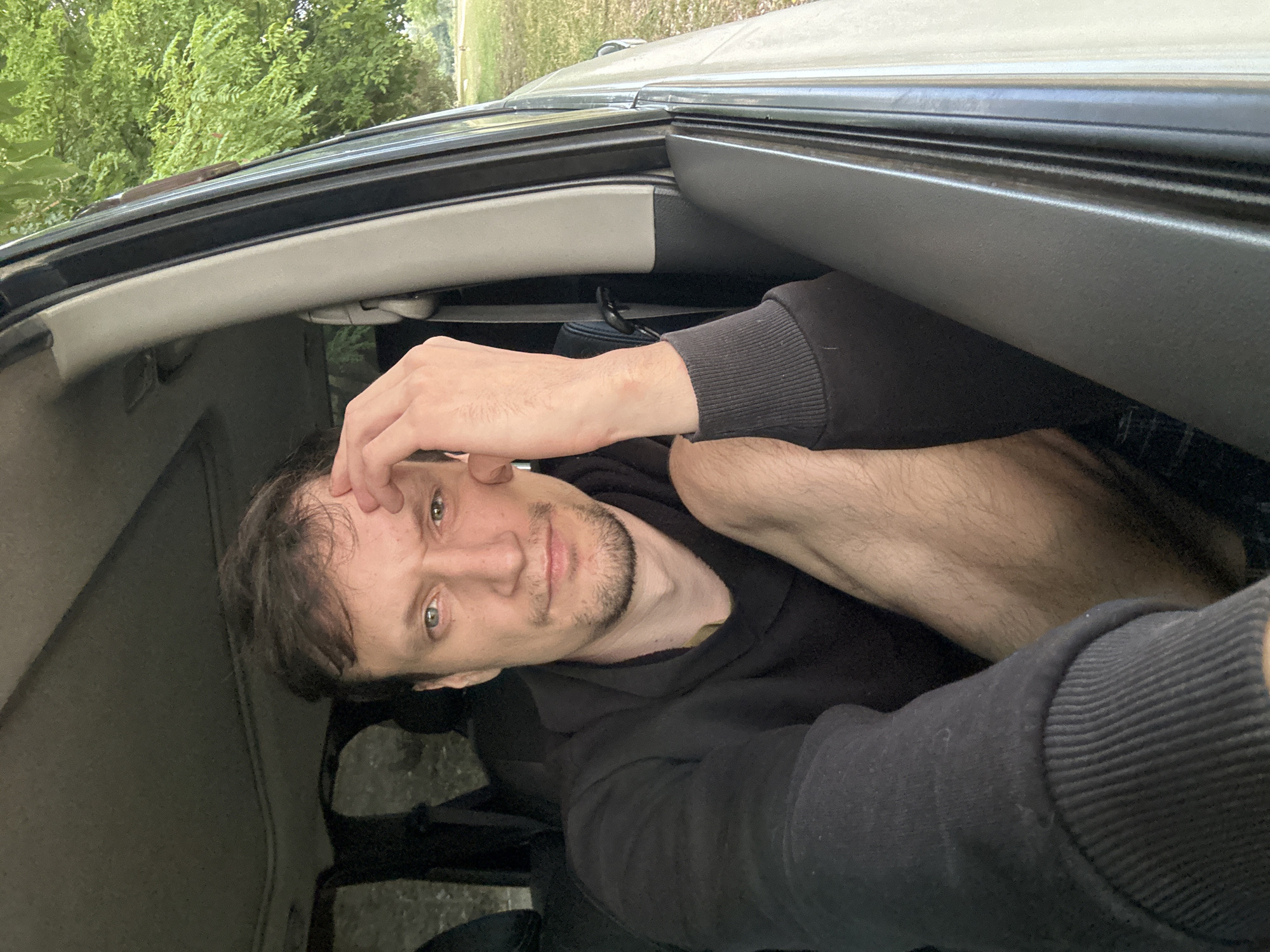 A man is sitting in the driver's seat of a car, looking directly at the camera. He has short, brown hair and a light beard. He is wearing a black hoodie. His right hand is resting on his forehead, and his left arm is resting on his leg. The car is parked outdoors, with trees and a grassy area visible through the open car door and window. The background suggests a rural or park-like setting outside of Makiv, Ukraine.