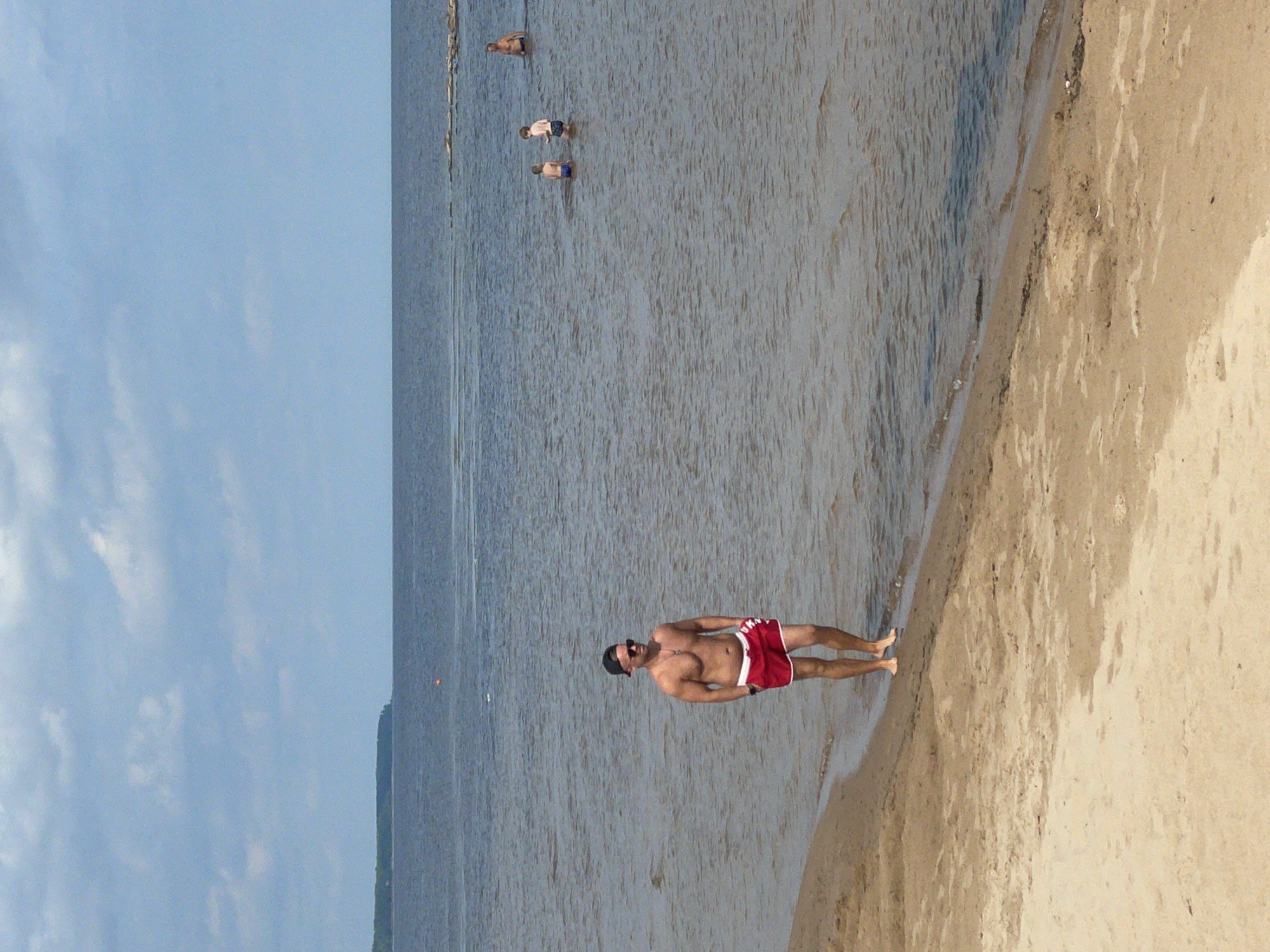 A man is standing in shallow water on the sandy shore of Lake Ladoga, Ladozhskoe Ozero, Russia. He is wearing red swim trunks, sunglasses, and a black baseball cap. His body is partially submerged in the water, and he appears to be walking towards the left of the frame. In the background, several other people are visible in the water, including two children and an adult man. The sky is overcast with scattered clouds, and the water is calm. The shoreline is a mixture of sand and some debris.