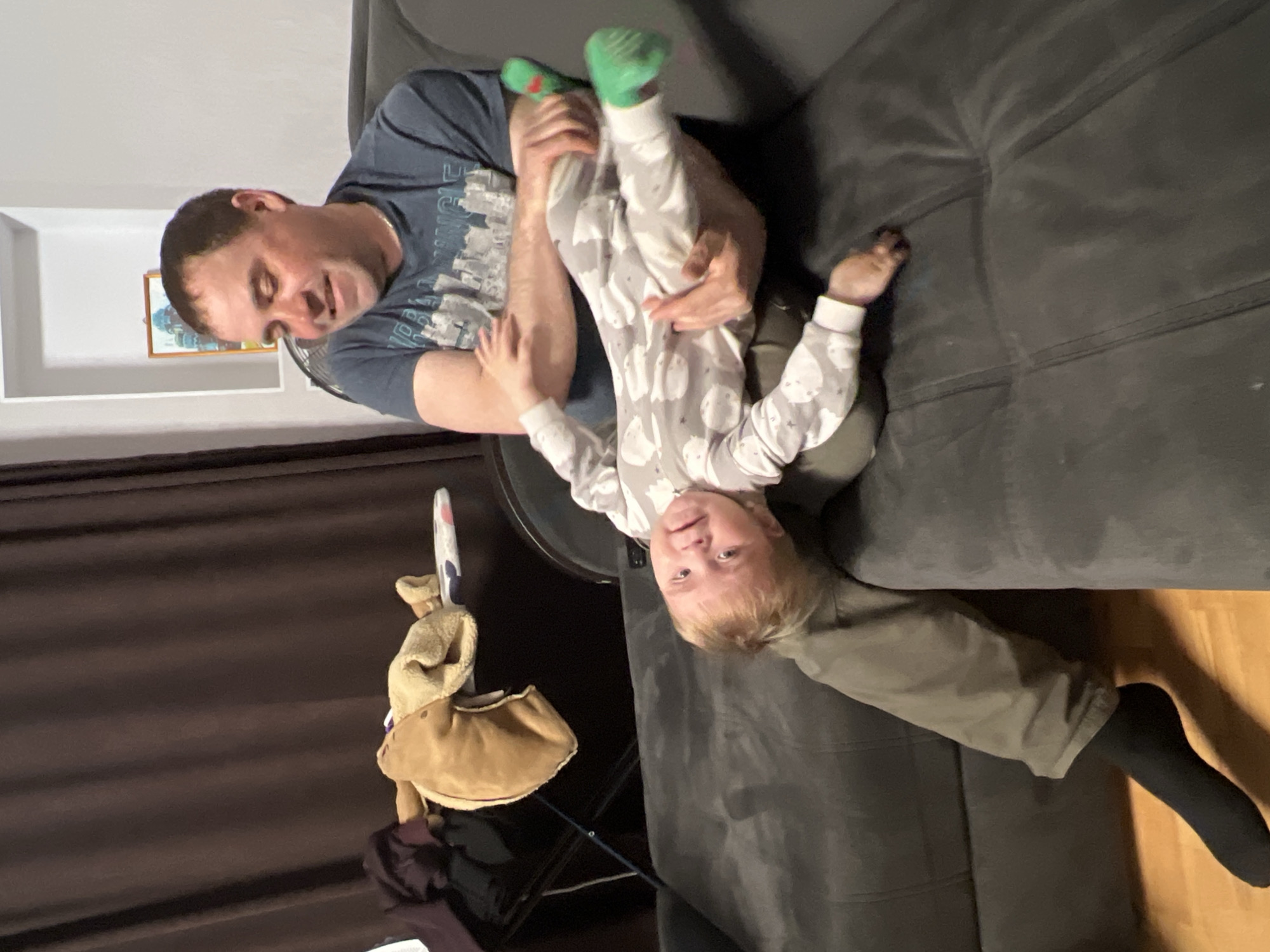 The image depicts an adult male playfully holding a young child upside down in an indoor setting, likely a living room in Perm, Russia. The man, with short brown hair, is seated on a dark grey sofa, wearing a dark blue t-shirt with an unreadable grey graphic design. He holds the child's legs, looking down with a gentle smile.
The child, appearing to be a toddler with light blonde hair, is dressed in a light grey full-body pajama suit featuring white ghost-like patterns and bright green socks. The child is looking directly at the camera with a slight, possibly mischievous, smile or smirk, evidently enjoying the upside-down position.
The background includes a white wall with a white shelf holding a small framed picture with blue and yellow hues. A dark brown curtain or fabric hangs on the left. A light brown plush toy with a colorful stick attachment is visible near the bottom left corner. Part of a wooden floor is seen on the right. The overall atmosphere is one of joyful familial interaction, characterized by playfulness and affection. The lighting appears to be artificial, suggesting the image was taken indoors, possibly in the evening.