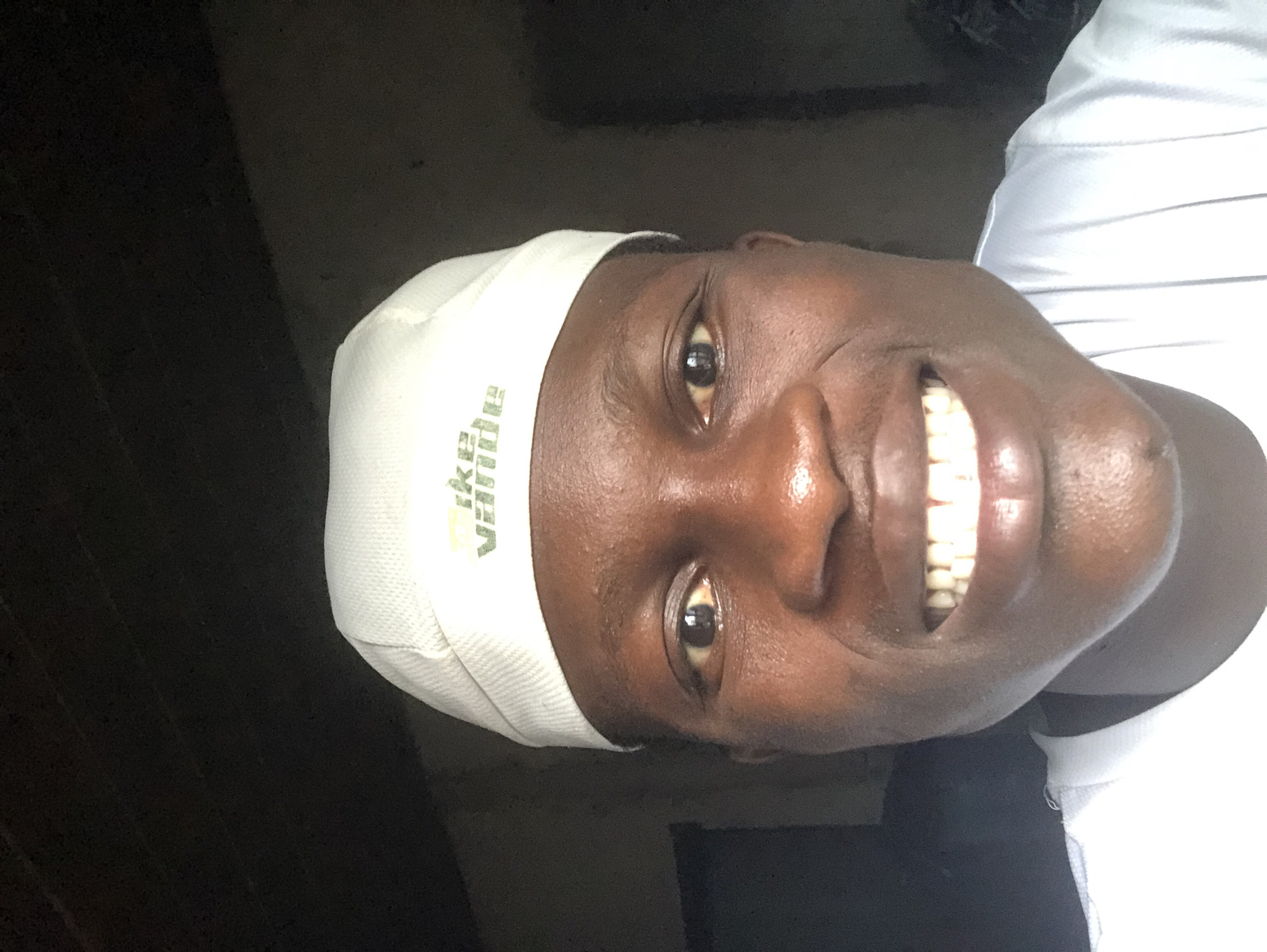 A man wearing a white cap and a white shirt is taking a selfie. The cap has a green logo. The man has dark skin and is smiling broadly, showing his teeth. The background is dark and appears to be indoors, possibly a room with a window. The lighting suggests it is daytime.