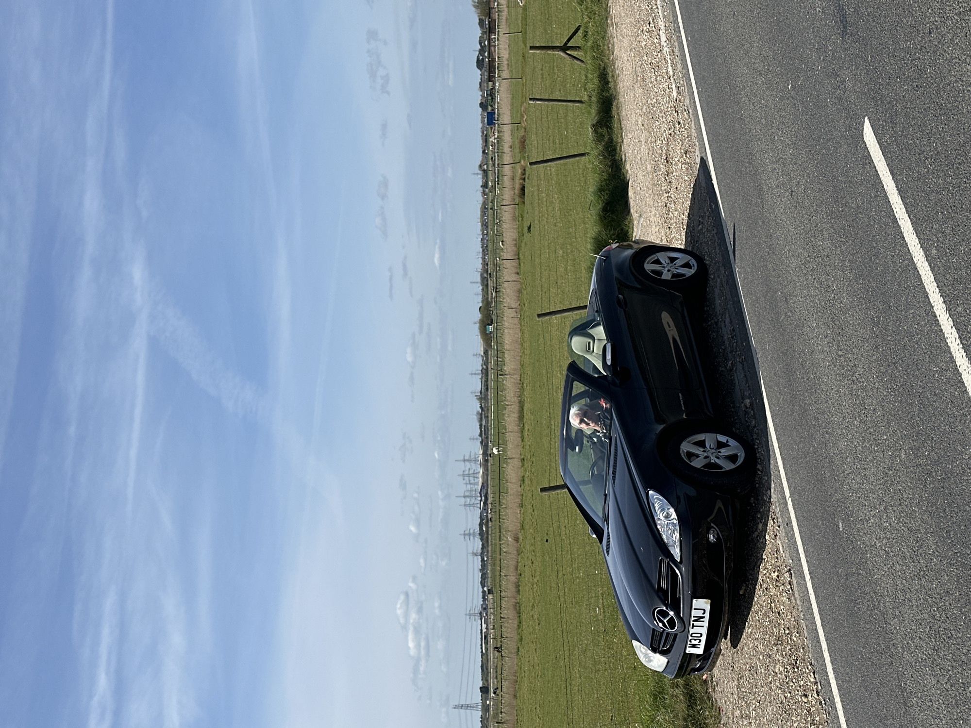 A black Mercedes-Benz SLK convertible is parked on the roadside in a rural area. The car faces towards the right of the frame. It is parked on the edge of an asphalt road with a white dividing line. Beyond the road is a gravel verge, and then a grassy field. A wooden post-and-wire fence runs through the field. In the distance, there are electrical transmission towers. The sky is light blue with wispy clouds. The location is in the Folkestone and Hythe District, United Kingdom. A person is visible in the driver's seat of the car. The license plate of the car reads "M30 TNJ".