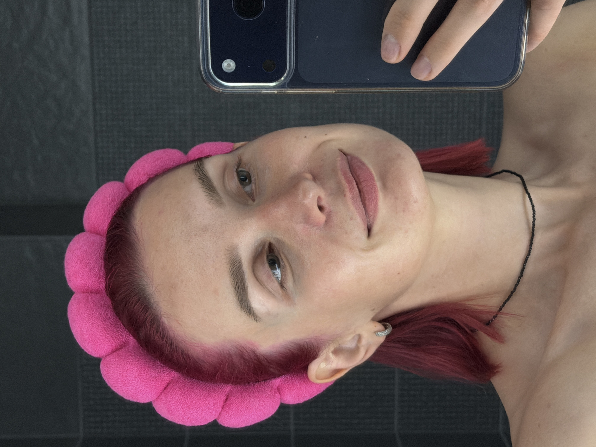 A young woman with pink hair, wearing a pink fluffy headband and a black beaded necklace, is taking a selfie in a bathroom. She is looking at her phone, which she is holding in her right hand. Her left arm is visible. The background appears to be a tiled wall. The image is a close-up of her face and upper body. There is no indication of the city of Chaikovsky, Russia in the image.