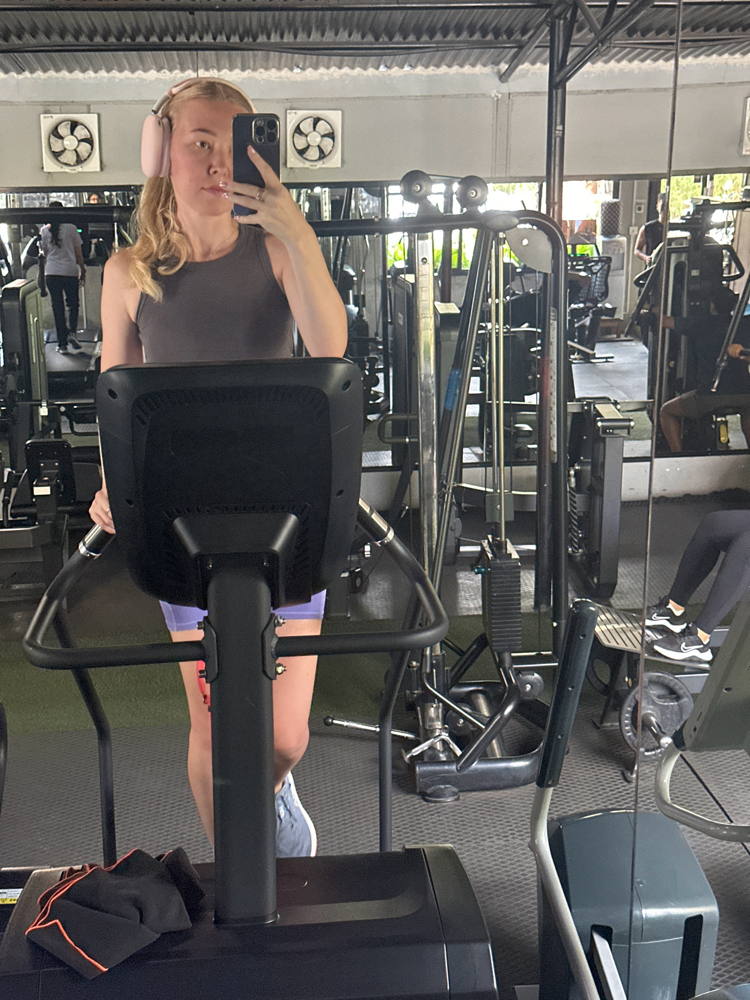 In the city of Lalitpur, Nepal, a woman with blonde hair and pink headphones is taking a mirror selfie in a gym. She is wearing a grey tank top and purple shorts. She is holding a black smartphone in her right hand and a ring is visible on her left ring finger. In the background, other gym equipment and people can be seen, including a treadmill and a weight machine. The gym appears to be indoors, with a corrugated metal ceiling and large windows reflecting the outdoors. The overall scene depicts a typical gym environment.