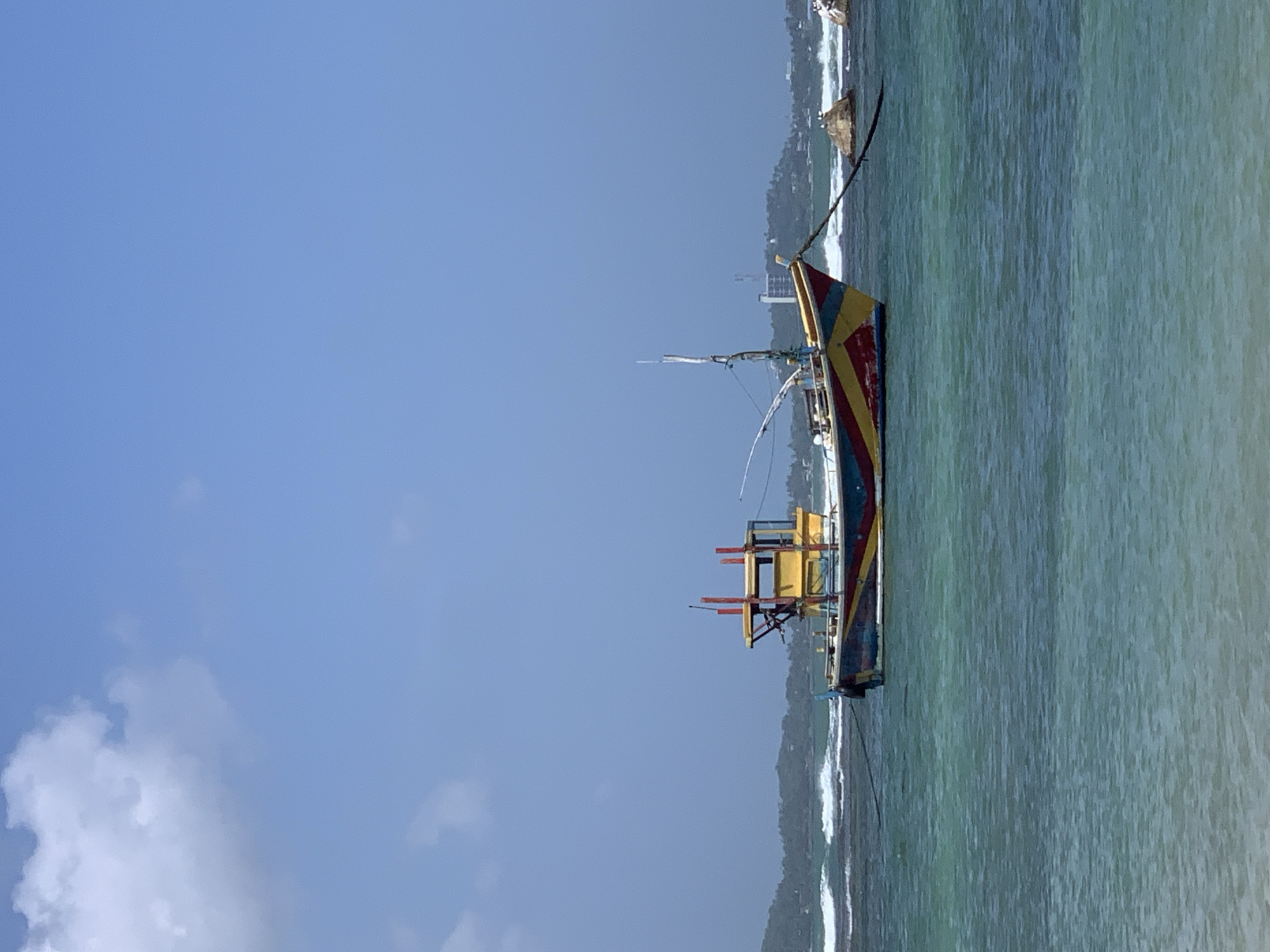A brightly colored fishing boat is moored in the turquoise waters off the coast of Weligama, Sri Lanka. The boat, painted with bold stripes of red, yellow, and blue, features a prominent yellow cabin structure with red accents. A dark rope extends from the boat's bow to an unseen anchor point. In the background, waves crash against the shore, and a lush, green coastline with buildings can be seen under a clear blue sky with scattered white clouds. The water is clear and shows ripples, suggesting a gentle current. The scene captures a typical day in this popular Sri Lankan coastal town.