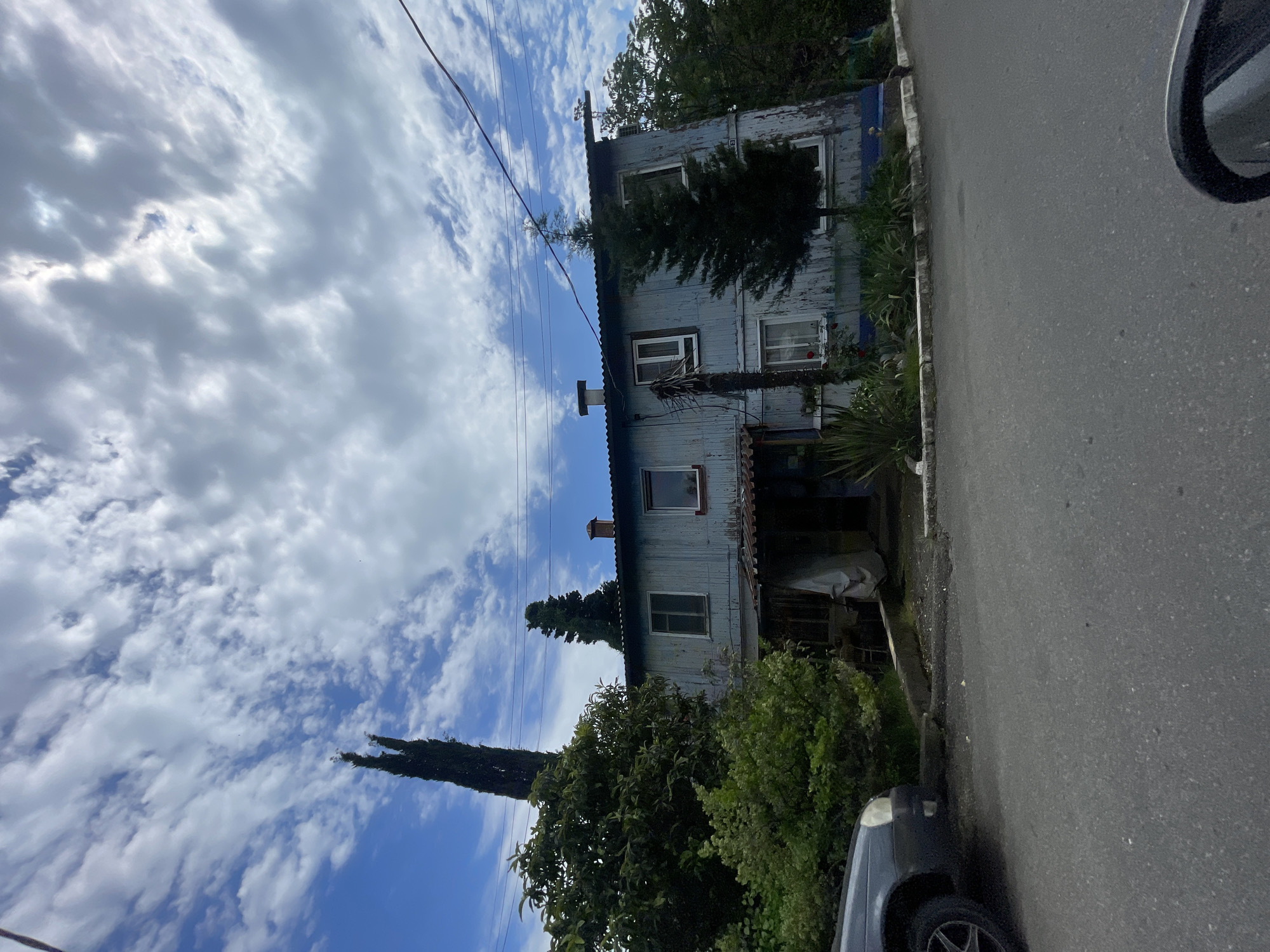 A two-story house with a corrugated metal roof and light blue, weathered wood siding is situated along a narrow asphalt road. The house has multiple windows with white frames. A compact dark car is partially visible parked on the roadside in front of the house, obscured by foliage. The cloudy sky overhead is partially obscured by trees. The image appears to be taken on a sunny day with scattered clouds. The location is presumed to be Sochi, Russia, as per the prompt's instructions.