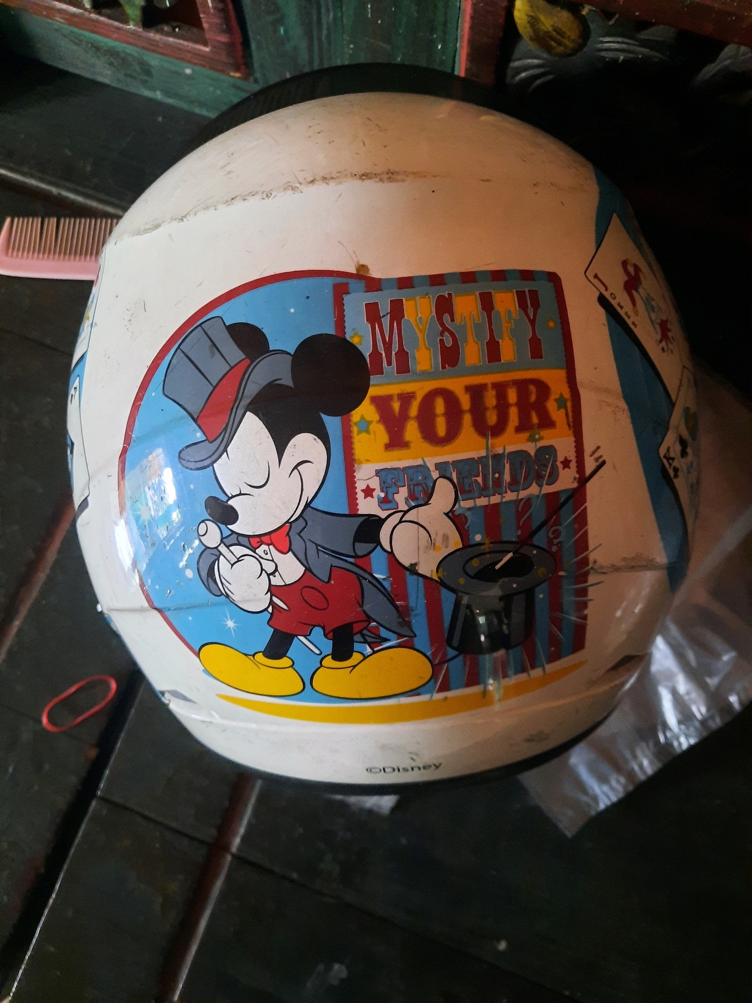 An overhead, slightly angled shot shows a white helmet with a Mickey Mouse sticker on its side. The sticker depicts Mickey Mouse in a magician's outfit, complete with a top hat and a magic wand. The words "MYSTIFY YOUR FRIENDS" are also visible on the sticker. The helmet is resting on a dark wooden surface, possibly a table or floor, with some reddish-brown and green wooden elements visible in the background. A pink comb and a red rubber band are also present on the wooden surface near the helmet. The overall scene appears to be indoors, possibly in a home or workshop setting, and is located in East Jakarta, Indonesia.