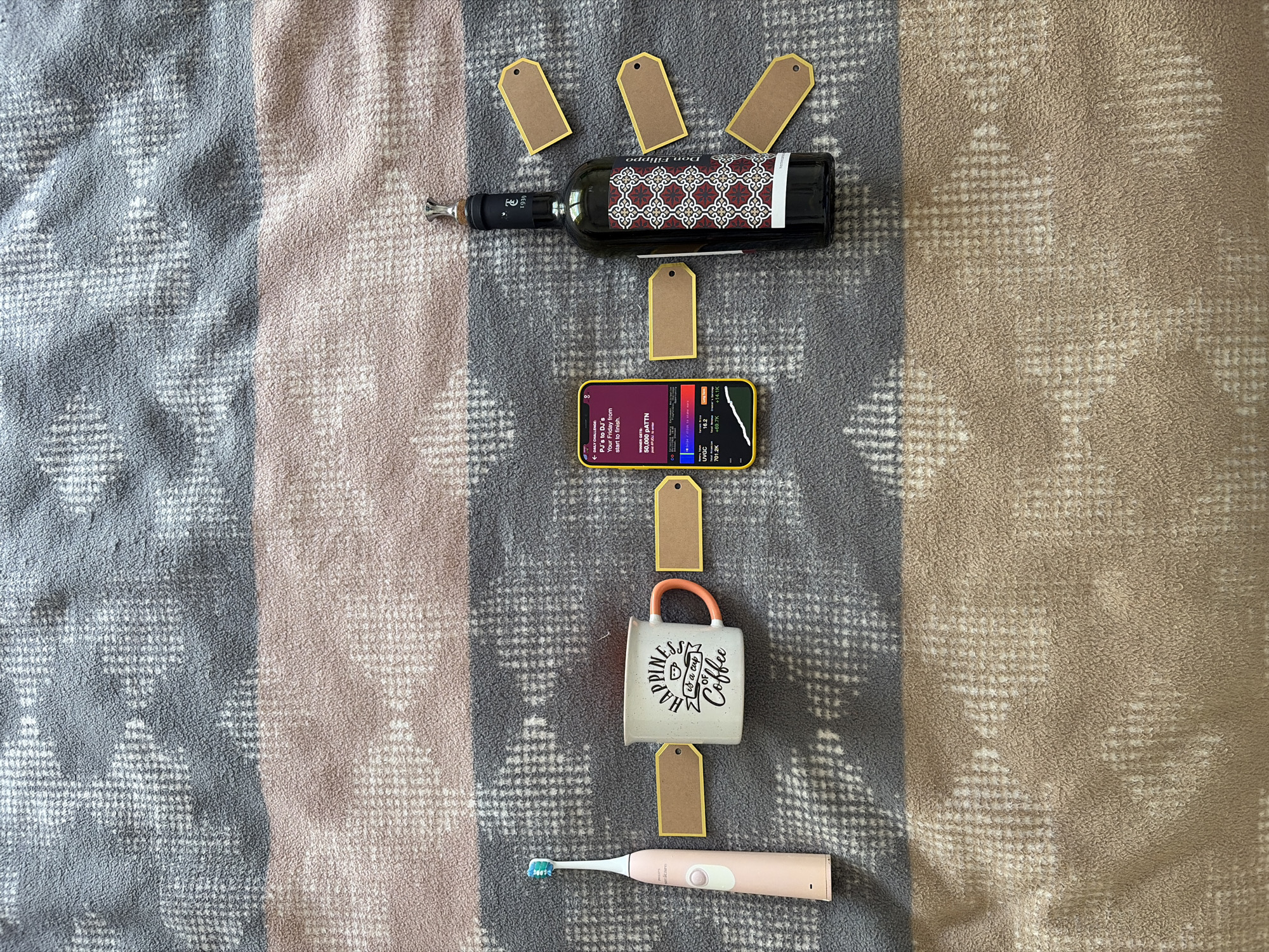 An overhead shot displays various objects arranged vertically on a patterned blanket, set in Budva, Montenegro. The blanket features wide stripes of grey, light brown, and pinkish-beige, with a subtle geometric design.
At the top, a dark glass wine bottle, labeled "Don Filippo" and "1939" with an intricate red and white pattern, is fitted with a silver pourer. Surrounding it are four blank, golden-edged brown tags, with another tag positioned below the bottle.
Next in line is a yellow-cased smartphone displaying an active screen. Visible text includes "DAILY CHALLENGE," "PJ's to DJ's," and "Your Friday from start to finish." Below this, "WINNER GETS: 50,000 PATTN" is shown, along with financial-looking figures like "UVGC 16.2," "701.2K," "+69.7K," and "+14.1K." Another blank tag sits beneath the phone.
Following the phone is a white ceramic mug with an orange handle, bearing the phrase "HAPPINESS is a cup of Coffee" in black text. Below the mug is another blank tag.
At the bottom, a pink Philips Sonicare electric toothbrush with a blue and white brush head completes the arrangement.
The objects, from wine to coffee to toothbrush, symbolically represent a progression of daily activities or a lifestyle, possibly linked to the "PJ's to DJ's" challenge on the phone, suggesting a transition from relaxation to social engagement throughout a day. The overall setup, with the neatly arranged items and blank tags, hints at a curated display or a collection of personal items for different moments of the day.