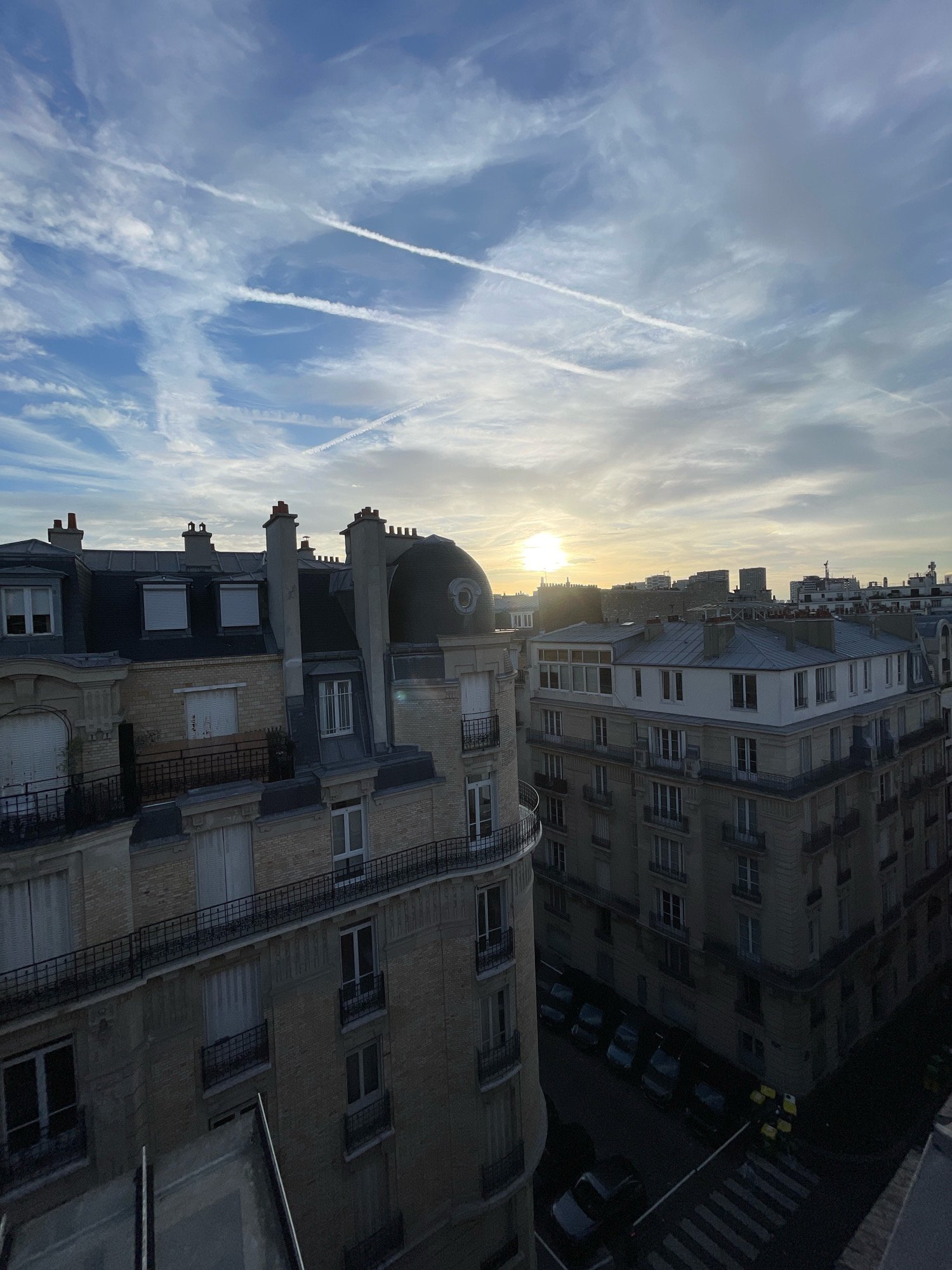 The image captures a view of Paris, France, during a sunrise or sunset, with the sun visible low on the horizon behind a hazy sky filled with wispy clouds and contrails. The foreground is dominated by the rooftops of Parisian buildings, characterized by their mansard roofs, chimneys, and dormer windows. The architecture suggests an older, classic Parisian style. In the middle ground, more buildings of a similar architectural style are visible, with balconies and multiple windows. The background reveals a skyline of taller, more modern buildings, indicating a mix of urban development. The street level, visible in the lower right, shows parked cars and a crosswalk, suggesting a residential or mixed-use urban environment. There are no discernible people or specific actions taking place in the scene. The overall impression is a quiet, urban landscape transitioning between day and night.