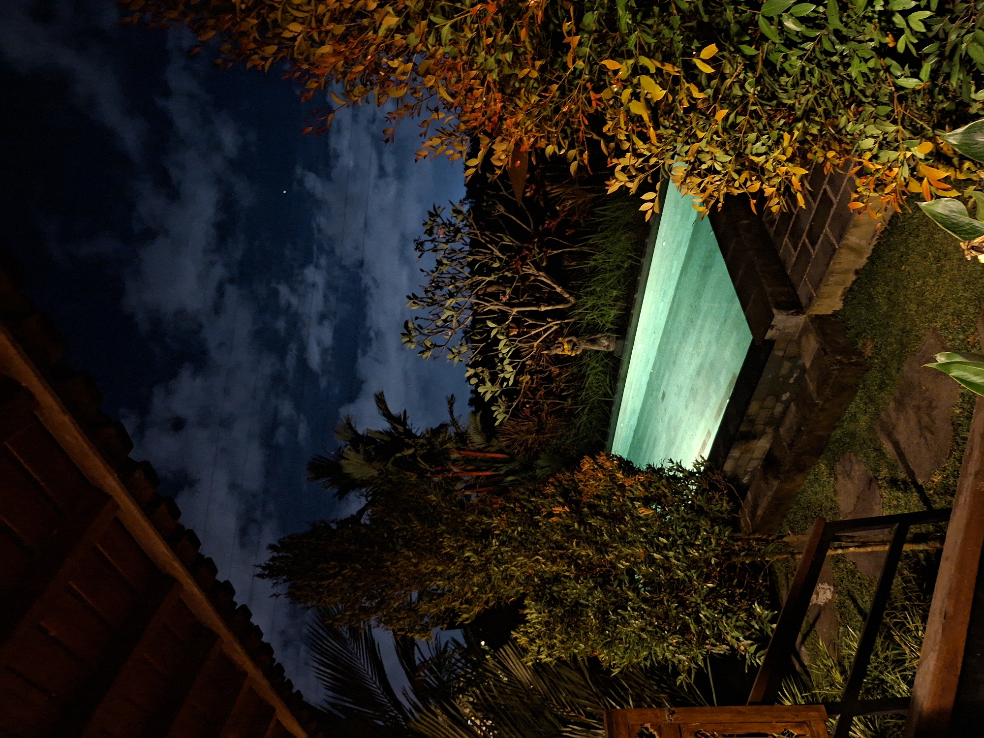 This vertical nighttime photograph captures a serene outdoor scene, likely a garden or courtyard within a resort or villa in a tropical location, consistent with Ubud, Indonesia. The central focus is a rectangular swimming pool, prominently illuminated with a vibrant turquoise glow, reflecting the dark surroundings.
The image is rich with lush vegetation. The upper frame is filled with dense foliage, its leaves displaying a range of colors from deep green to warm yellows and oranges, suggesting they are lit by artificial lights. Below this, various tropical trees, including visible palm fronds and a bare-branched tree resembling a frangipani, are silhouetted against the night sky. The ground around the pool features a mix of green grass and stone pavers.
The sky is a deep, dark blue, adorned with soft, illuminated clouds, and a tiny, distant star or light is visible. To the far left, the warm-toned wooden roofline and eaves of a building are partially visible, hinting at traditional architecture. In the lower right foreground, a dark wooden railing or fence provides structural detail.
The time of day is clearly night, emphasized by the dark sky and the artificial illumination of the pool and some of the foliage. No people are visible in the scene, and there is no discernible text. The overall impression is one of tranquility and exotic ambiance, characteristic of a peaceful evening in a tropical setting.