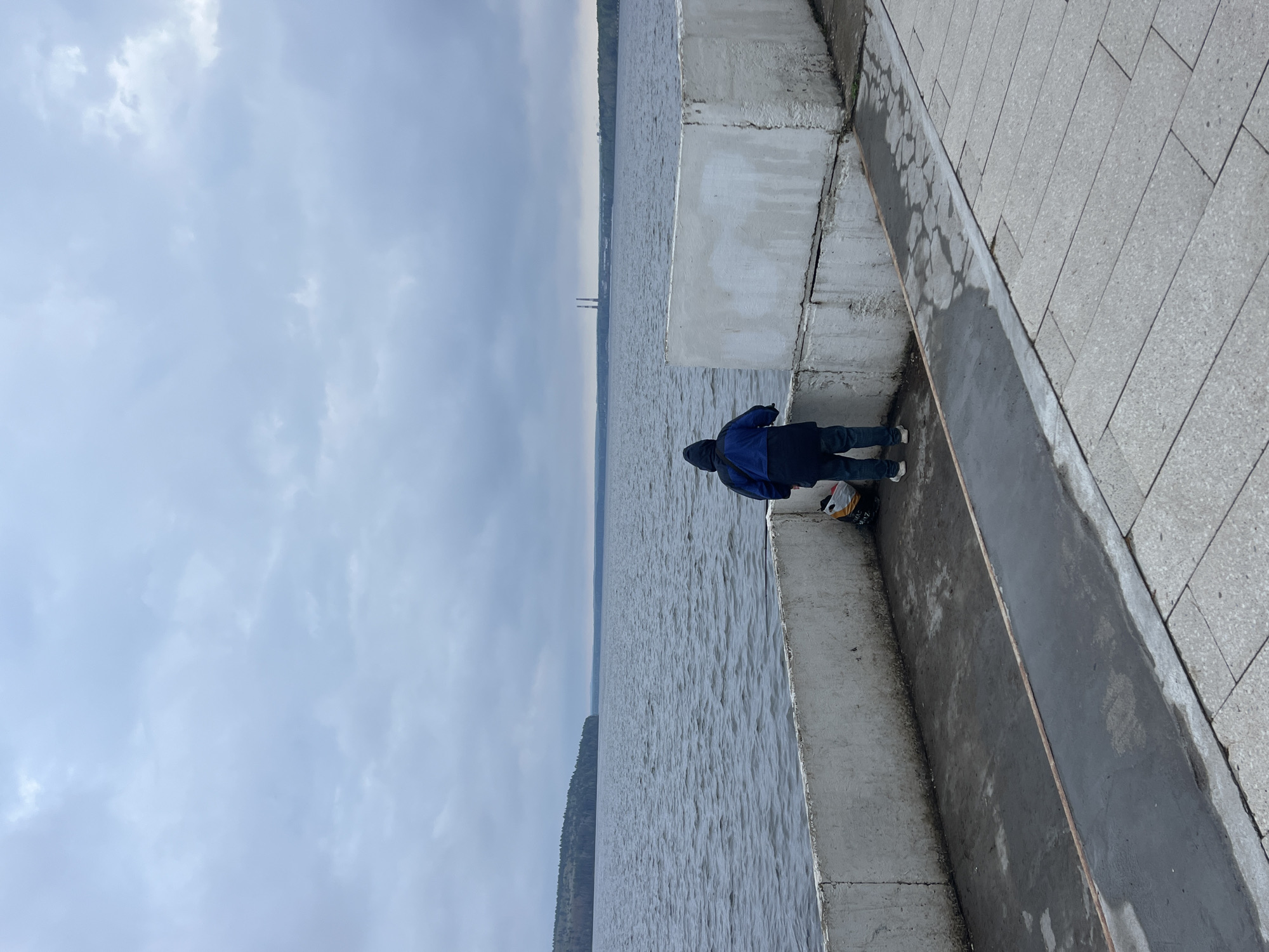 A person in a dark blue jacket and dark pants stands at the edge of a concrete embankment, looking out over the water. The water is gray and appears to be a large body of water like a river or a lake. In the distance, the opposite shoreline is visible, with a dark treeline along the top. Above, the sky is overcast with gray clouds. The embankment is made of concrete blocks and leads to a paved walkway made of gray stone tiles. A bucket sits on the walkway next to the person. The location is likely the city of Chaikovsky, Russia.