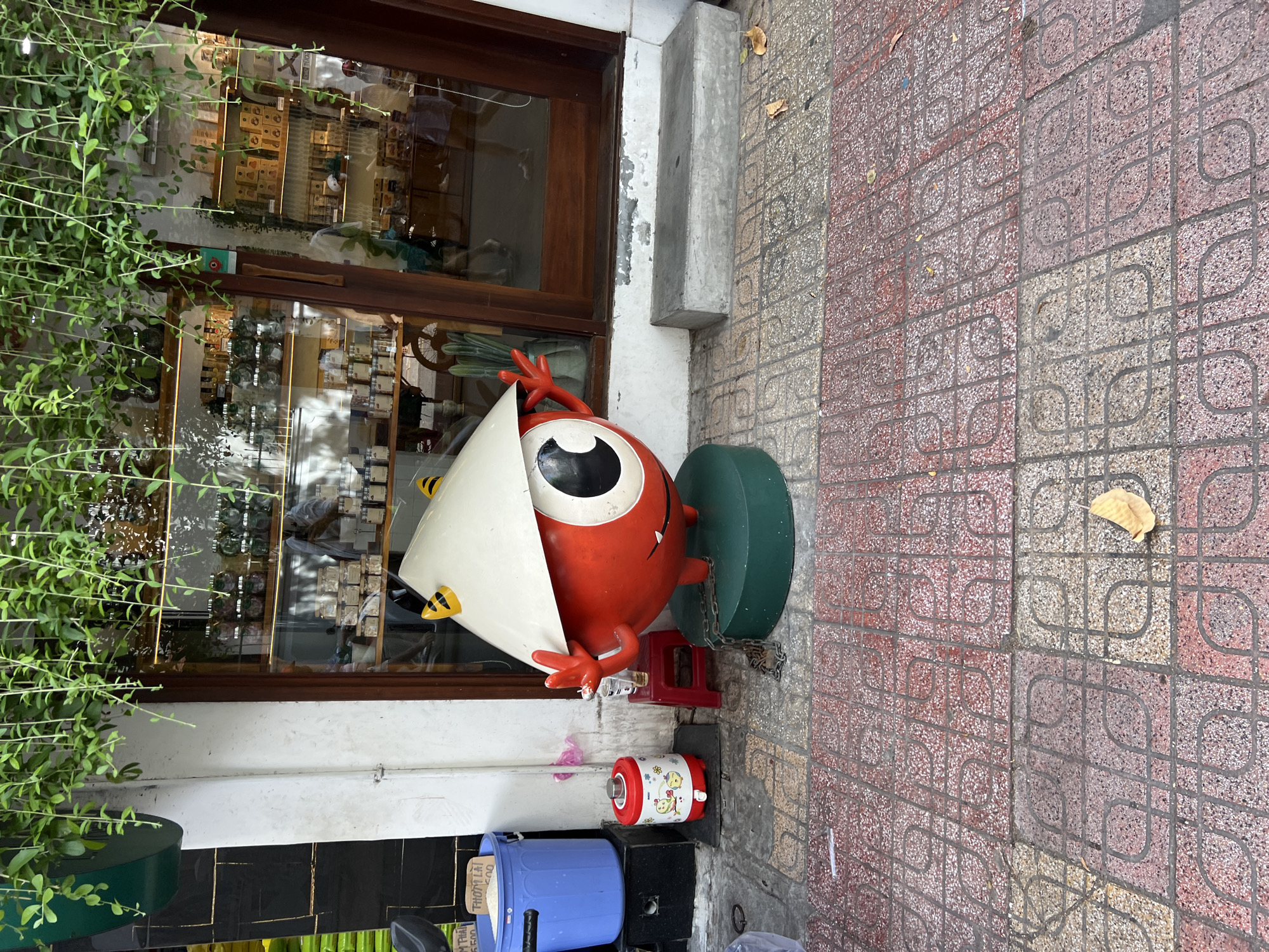 The image shows a statue of a red, round character wearing a conical hat and yellow horns, standing on a green platform in front of a store. The statue is chained to the platform with a metal chain. To the left of the statue, there is a blue trash can and a small red water dispenser. The store has a large glass window displaying shelves filled with jars and boxes. Above the store, green vines hang down. The ground in front of the store is paved with red and beige patterned tiles. A few dry leaves are scattered on the tiles. The scene appears to be on a street in Nha Trang, Vietnam, as suggested by the conical hat worn by the statue, which is a traditional Vietnamese hat.