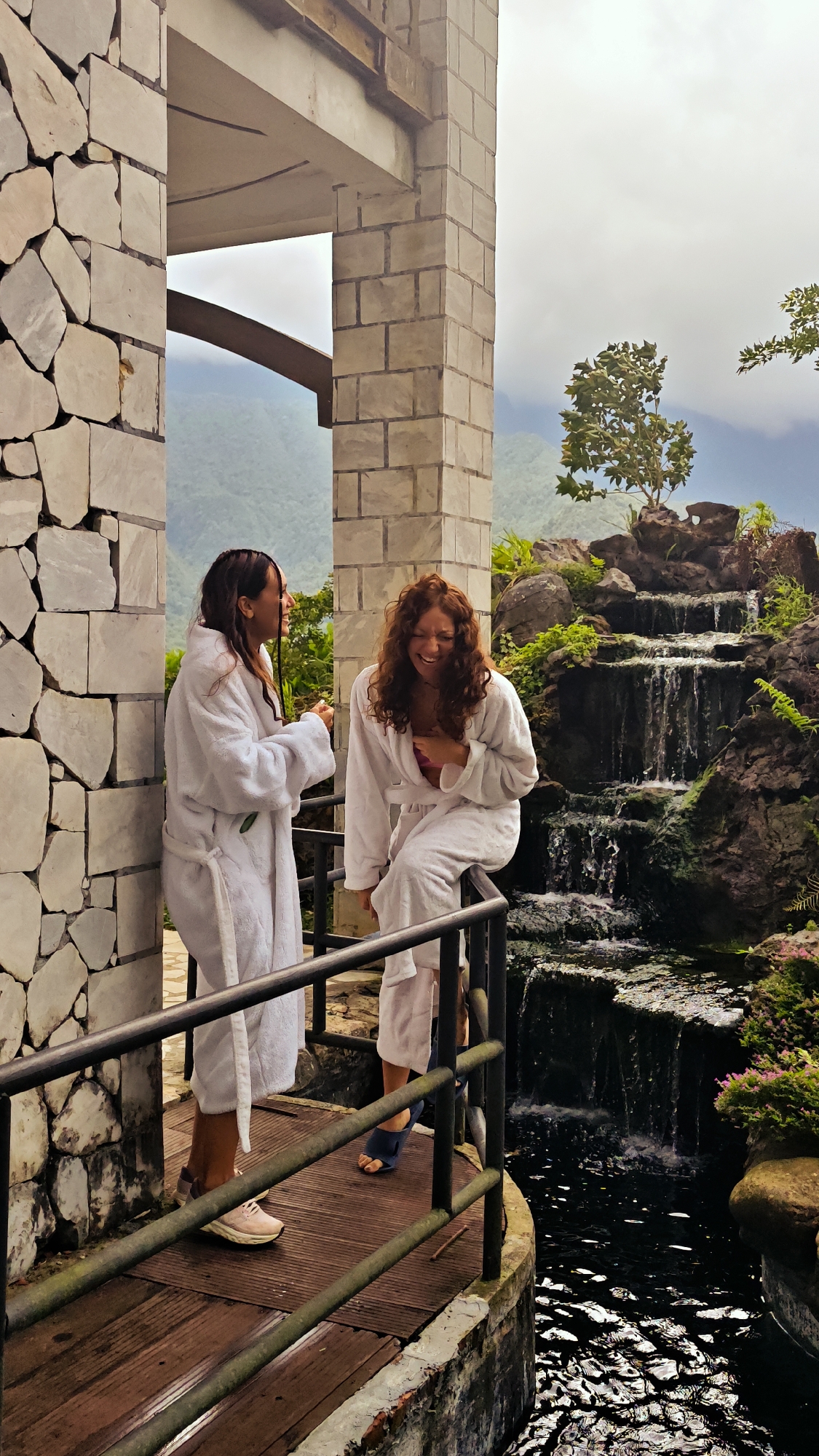 Two women wearing white bathrobes stand on a wooden platform enclosed by a black metal railing in Cat Cat Village, Vietnam. The woman on the left has dark hair and is facing the woman on the right, who has red curly hair and is perched on the railing. A stone structure flanks them on the left and a brick structure is behind the woman on the right. A waterfall cascades down rocks and greenery to a dark pool of water. Behind the waterfall, mountains are visible in the distance. The woman on the left wears tan sneakers, while the woman on the right wears a blue sandal.
