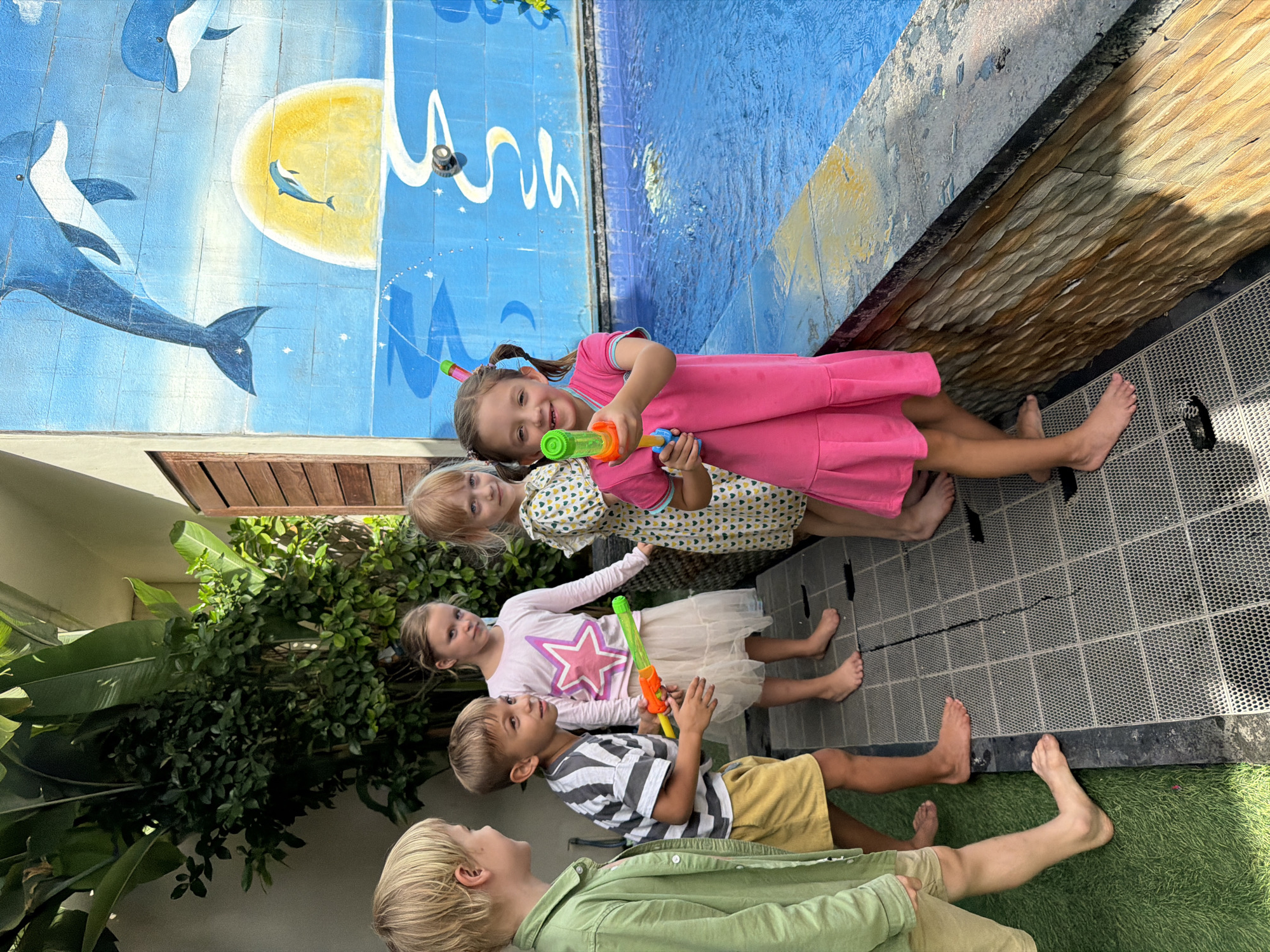 Five children are standing outdoors, near a swimming pool with a mural of dolphins on a blue wall behind them. The children are of varying ages and wear casual clothing. One child wears a pink dress and holds a water gun. Another child wears a patterned dress, and a third wears a pink shirt with a star. A fourth child wears a striped shirt and yellow shorts, and the last child wears a green shirt and khaki shorts. The ground is covered in grey tiles and artificial grass. The scene is in Kuta, Indonesia.
