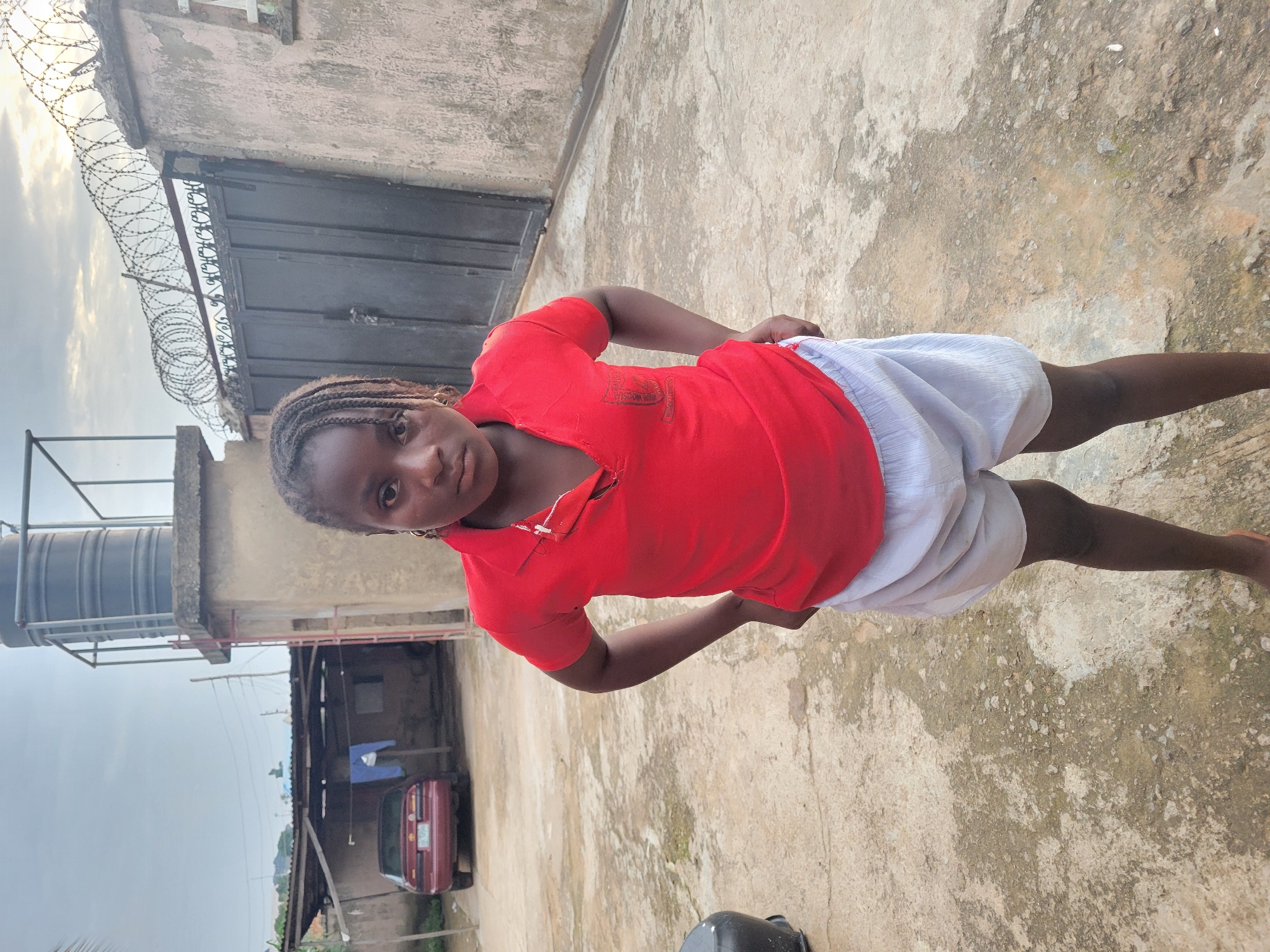 This is a vertical-format, full-body shot of a young Black person standing on a concrete surface in what appears to be an outdoor yard. The subject is wearing a red polo shirt and white shorts. Their hair is braided into cornrows. The person is looking directly at the camera with a neutral expression. The background shows a building with a metal gate topped with barbed wire, a large water tank on a metal frame, and a covered structure with a car parked underneath. A red car is visible parked under the covered structure. The sky is overcast, suggesting either early morning or late afternoon. The concrete ground is weathered and has some patches of discoloration. A black plastic bucket is visible in the lower right corner of the frame. The scene appears to be in a residential or compound area in Madalla, Nigeria. No visible text is present in the image.