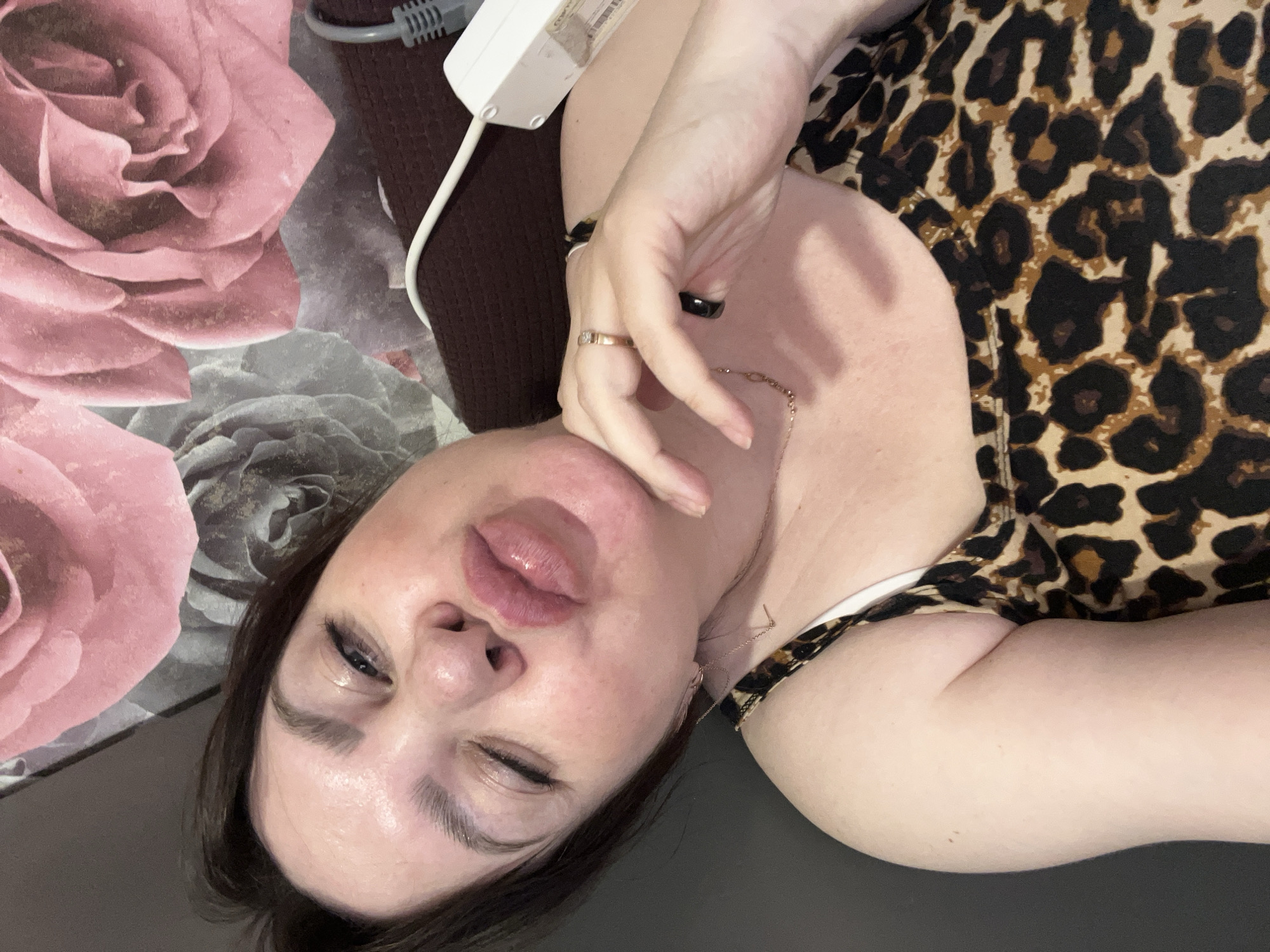 An adult female is captured in a close-up, angled portrait, likely a selfie, while lying down in an indoor setting in Noviy, Russia. She has dark hair and is wearing a leopard print top with a white strap visible on her left shoulder. Her face is turned towards the camera, with one eye winking or partially closed and her lips slightly puckered, conveying a playful or relaxed expression. Her skin appears fair, and her eyelashes and eyebrows are prominent.
Her right hand rests near her face, adorned with a gold-colored ring featuring a stone on her ring finger and a black ring on her middle finger. A delicate gold-colored chain necklace drapes around her neck. A white electronic device with a gray cable, possibly for personal care, is visible near her right shoulder.
The background consists of bedding or a cushion patterned with large pink and gray roses, suggesting a comfortable, personal space like a bedroom or living room. The lighting is soft, illuminating her features without harsh shadows. No clearly decipherable text is visible on any objects.