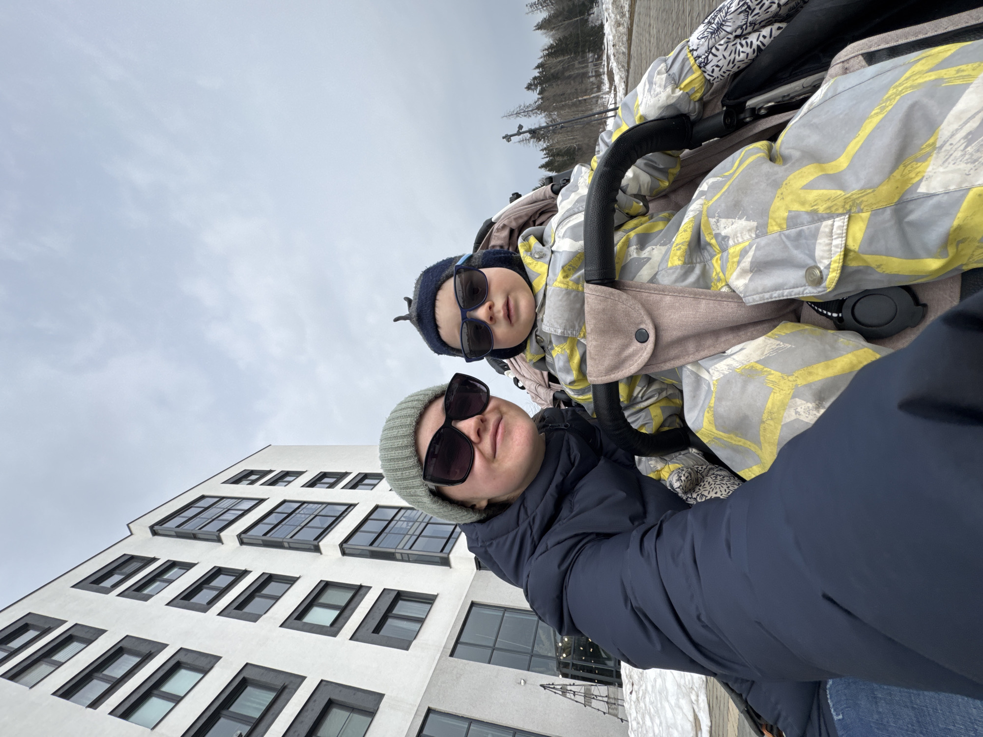 An outdoor scene in Izhevsk, Russia, depicts an adult female and a child on an overcast day. The female, positioned on the left and partially obscured from the chest down, wears a dark blue puffer jacket, a green knit beanie, and dark sunglasses. She holds the camera for a selfie, looking directly into it with a slight smile. To her right, a child is seated in a beige stroller, dressed in a grey and yellow patterned winter suit, a dark blue knit beanie, and dark sunglasses, also looking towards the camera with a neutral expression. In the background on the left, a modern multi-story building with a white facade and prominent dark grey window frames and vertical accents rises. Patches of melting snow are visible on the paved ground in the foreground and mid-ground. Further back on the right, a line of bare trees and conifers stands against the light grey sky.