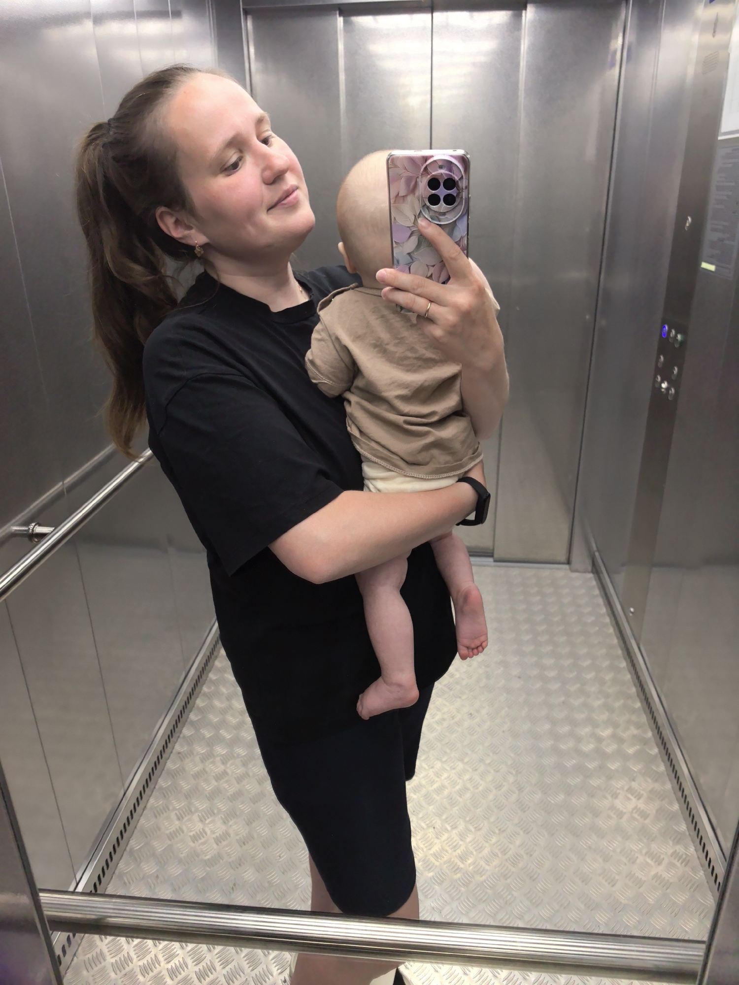 A woman is holding a baby in an elevator, taking a selfie with her smartphone. The woman is wearing a black t-shirt and shorts, her brown hair is pulled back in a ponytail. She is smiling at the baby, who is dressed in a tan shirt and light-colored diaper. The woman is wearing a smartwatch on her left wrist. The elevator interior is brushed metal. The floor is a textured silver metal. A handrail is visible along the left wall of the elevator. The elevator control panel is visible on the right wall. The location is Unknown, Unknown.