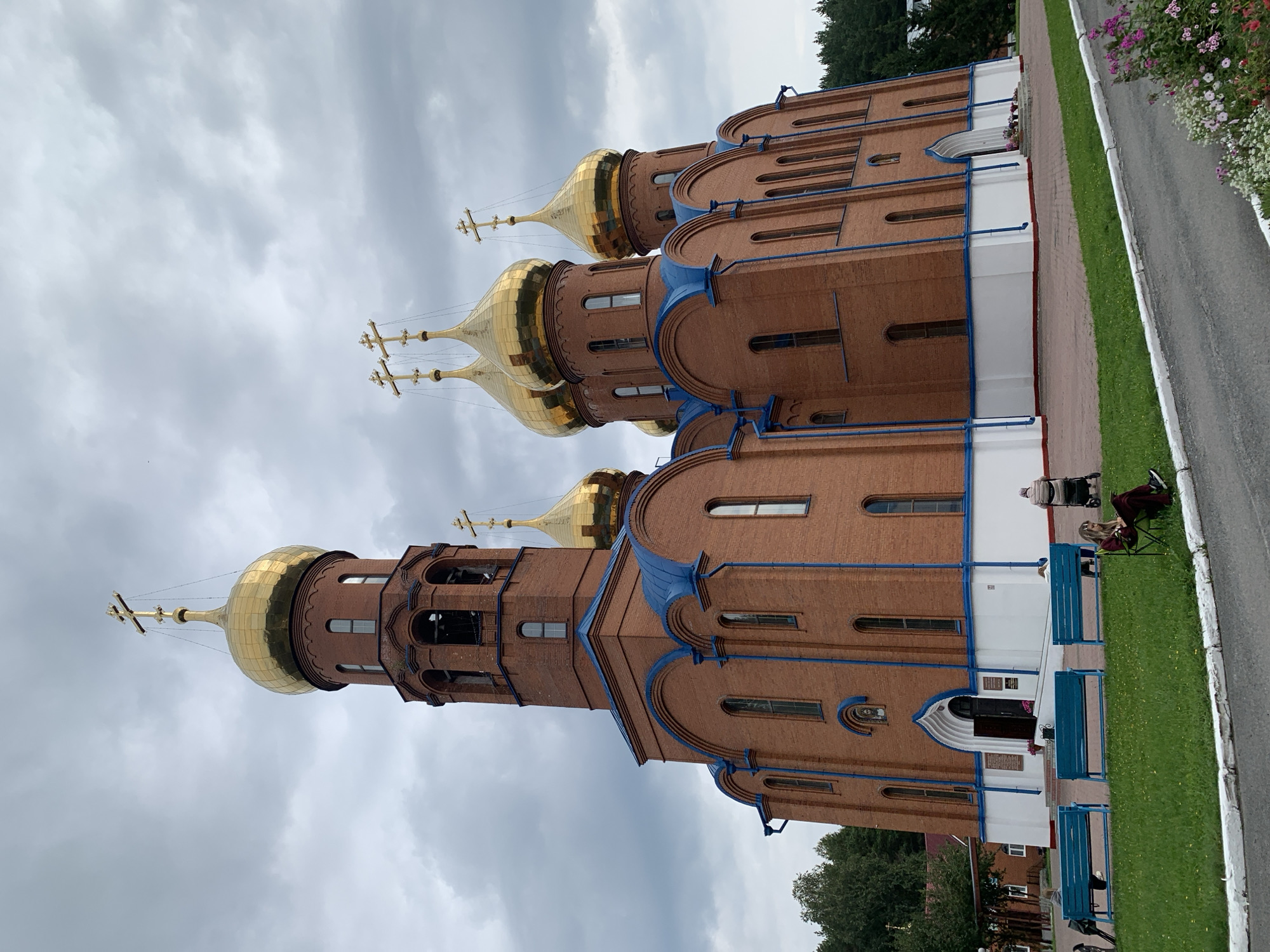 This image depicts the Church of the Intercession of the Most Holy Mother of God, located in the city of Prokopyevsk, Russia. The church is a prominent brick structure with a central bell tower and multiple golden domes topped with crosses. The exterior features blue trim along the rooflines and white detailing at the base. In front of the church, there are several blue benches situated on a grassy area, and a paved walkway leads to the entrance. On the right side of the walkway, a young woman sits in a chair, reading a book. Further back, an elderly person is pushing a stroller, and another individual is walking. The sky is overcast with dramatic clouds. The surrounding area appears to be natural with trees visible in the background.