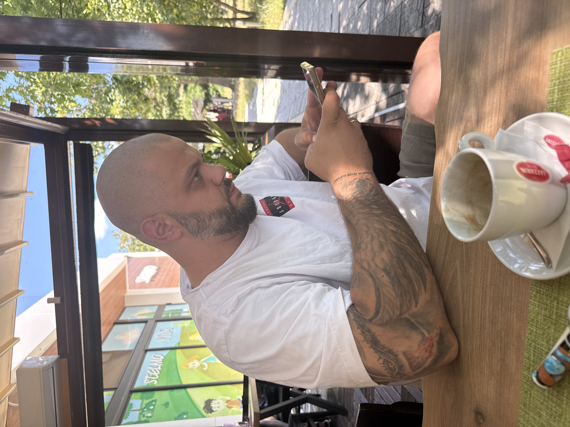 A man with a shaved head and beard is sitting at a wooden table in an outdoor cafe in Plovdiv, Bulgaria. He is wearing a white t-shirt and is looking down at his phone, which he is holding in his hands. His left arm is heavily tattooed. A white coffee cup and saucer are on the table in front of him. The background shows trees and a building with colorful murals.