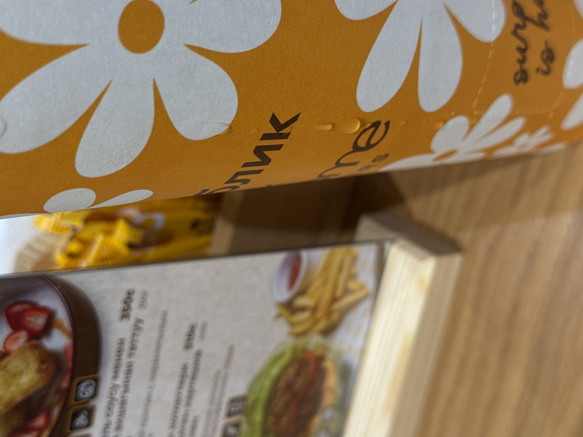 The image captures a close-up, slightly angled view of a table in what appears to be a cafe or restaurant setting. The most prominent object is a cylindrical container, likely a cup or bottle, adorned with a vibrant orange background and a pattern of white, daisy-like flowers. Several small water droplets are visible on its surface.
Partially visible text on the container includes a Cyrillic character "к" (appearing reversed due to the angle) and the English phrase "surprise is h..." in a handwritten style, indicating an English phrase possibly continuing as "surprise is here" or similar.
Below and to the left of the container is a menu or booklet, positioned at an angle. It features blurred images of food items. On the left side of the menu, there's an image of what looks like pancakes or french toast topped with strawberries. On the right side, there's a picture of a burger served with french fries and a small cup of sauce. The menu contains text written in a Cyrillic script. Decipherable words and numbers include "350с" and "010с" (likely prices), and the phrase "апельсиновый капучино" (orange cappuccino).
The objects are resting on a light brown, wooden surface, which appears to be a table or counter. In the blurred background, some yellow, indistinct objects are visible, possibly food packaging or other items. There are no people visible in the image. The scene suggests an indoor environment where food and drinks are served.
