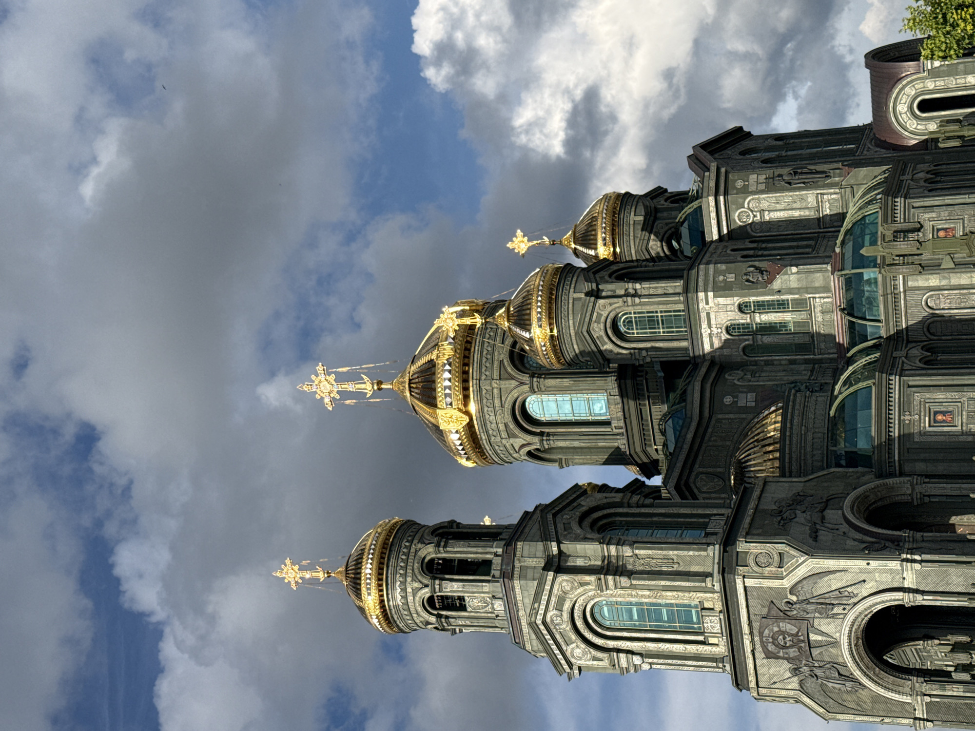 The image shows a large, ornate cathedral with multiple golden domes topped with crosses. The cathedral is made of dark gray stone and features intricate carvings and arched windows. The sky is cloudy with patches of blue. The cathedral is located in the city of Zelenaya Roshcha, Russia.