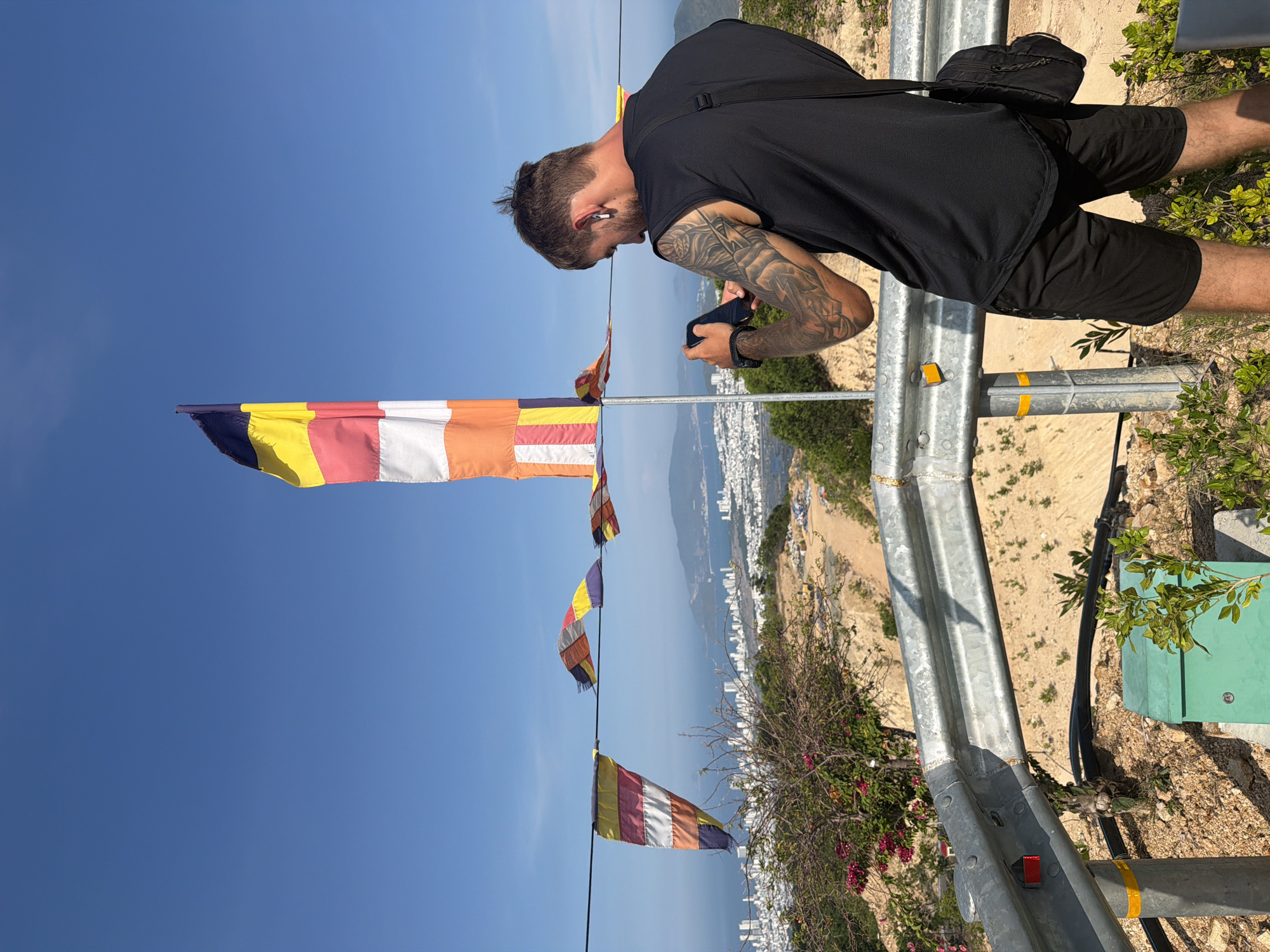 A man with a tattooed arm and a partially tied-up hairstyle is standing next to a guardrail on an elevated area in Nam Nha Trang Ward, Vietnam, looking down at his mobile phone. He's wearing a black sleeveless shirt and black shorts. A black bag hangs from his shoulder. Behind him and below, the cityscape of Nha Trang stretches to the coastline. In the foreground, a string of colorful prayer flags is stretched between poles. Vegetation and utility equipment are visible alongside the guardrail. The sky above is clear blue.