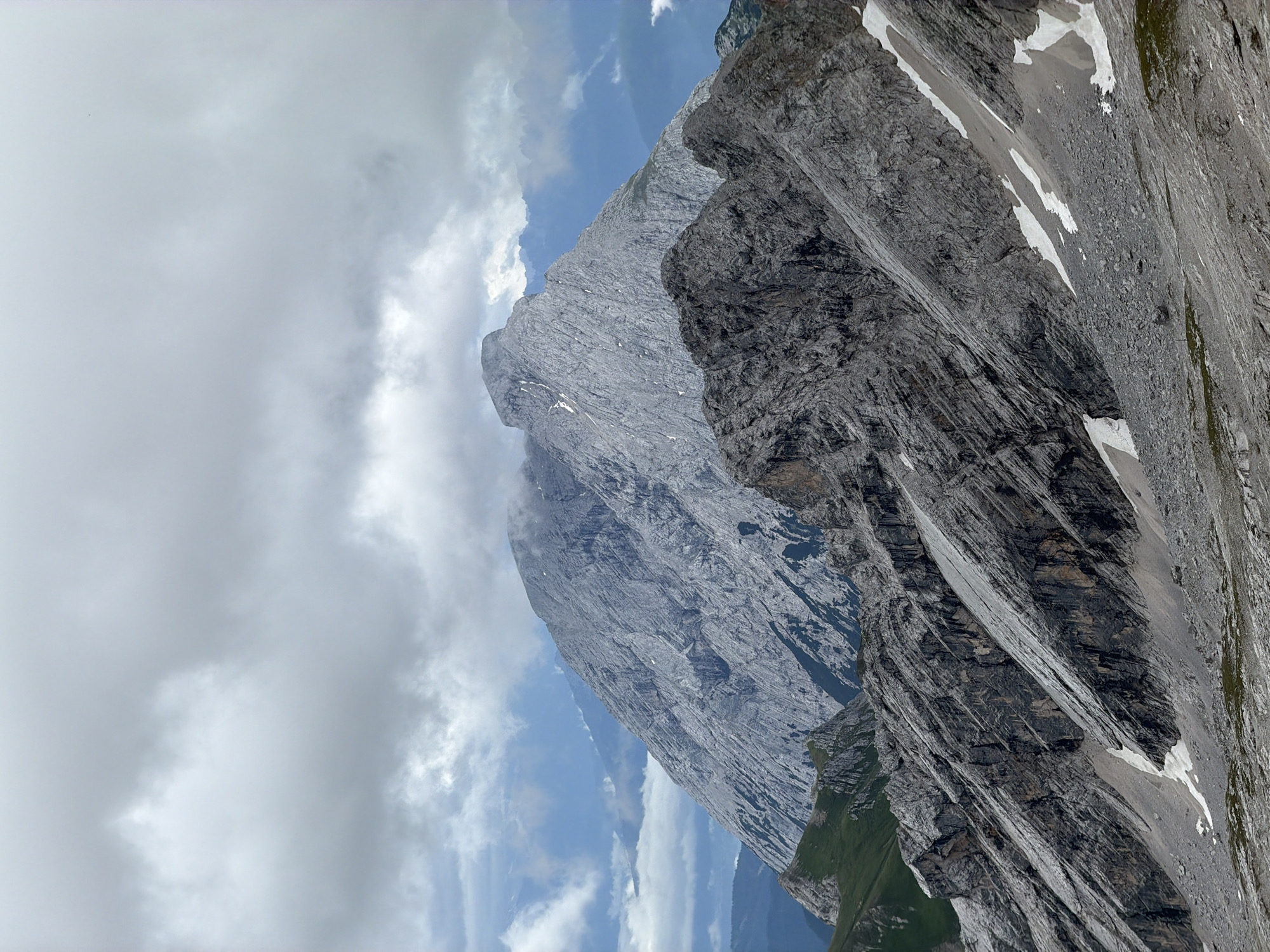 The image depicts a rugged mountain landscape under a cloudy sky. The foreground is dominated by steep, rocky slopes with patches of snow and sparse green vegetation. In the middle ground, a darker, more jagged mountain range rises, characterized by sharp ridges and deep crevices. The background features a massive, lighter-colored mountain with a prominent peak, partially shrouded in mist. Distant mountain ranges are visible in shades of blue and grey, indicating depth and atmospheric conditions. The overall scene conveys a sense of vastness and natural wilderness, typical of alpine environments. Given Garmisch-Partenkirchen's location in the Bavarian Alps, this scene is likely situated within the mountainous region surrounding the city.