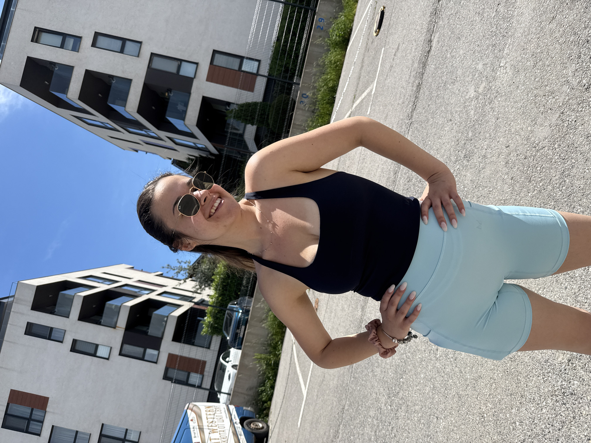 A woman stands in a paved parking lot in Sofia, Bulgaria. The lot is surrounded by a modern building with dark windows and a black metal fence. A blue van with Cyrillic text is visible in the background. The woman is wearing a dark tank top, light blue shorts, and sunglasses. She has her hands on her hips and is smiling at the camera. The sky is blue and sunny.