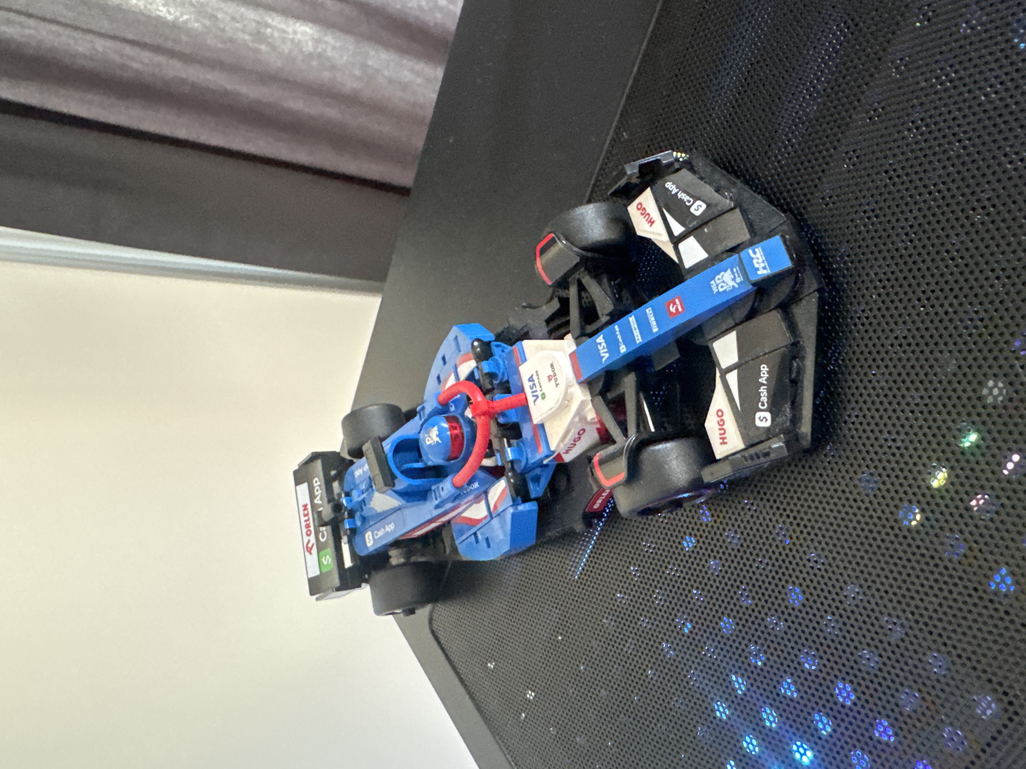 This is a photograph of a toy Formula 1 race car, likely a replica, placed on a black, perforated surface. The car is predominantly blue and white, with red accents. It features various sponsor logos printed on its body, including "ORLEN," "VISA," "Cash App," "HUGO," and "TUDOR." Some of these logos are partially obscured or appear as stickers. The car has black wheels and a visible cockpit area with red roll hoop detailing. The perforated surface it rests on has a mesh-like pattern and is illuminated by what appears to be colorful LED lights, suggesting it might be part of a computer case or a decorative item. In the background, a dark gray curtain and a light-colored wall are visible, providing a glimpse of an indoor setting. The lighting in the image is somewhat dim, with the colorful LEDs of the surface being the most prominent light source. The photograph seems to be taken at a slightly elevated angle, looking down at the toy car. There are no people visible in the image. The overall impression is of a display of a collectible item in a modern, possibly tech-oriented environment.