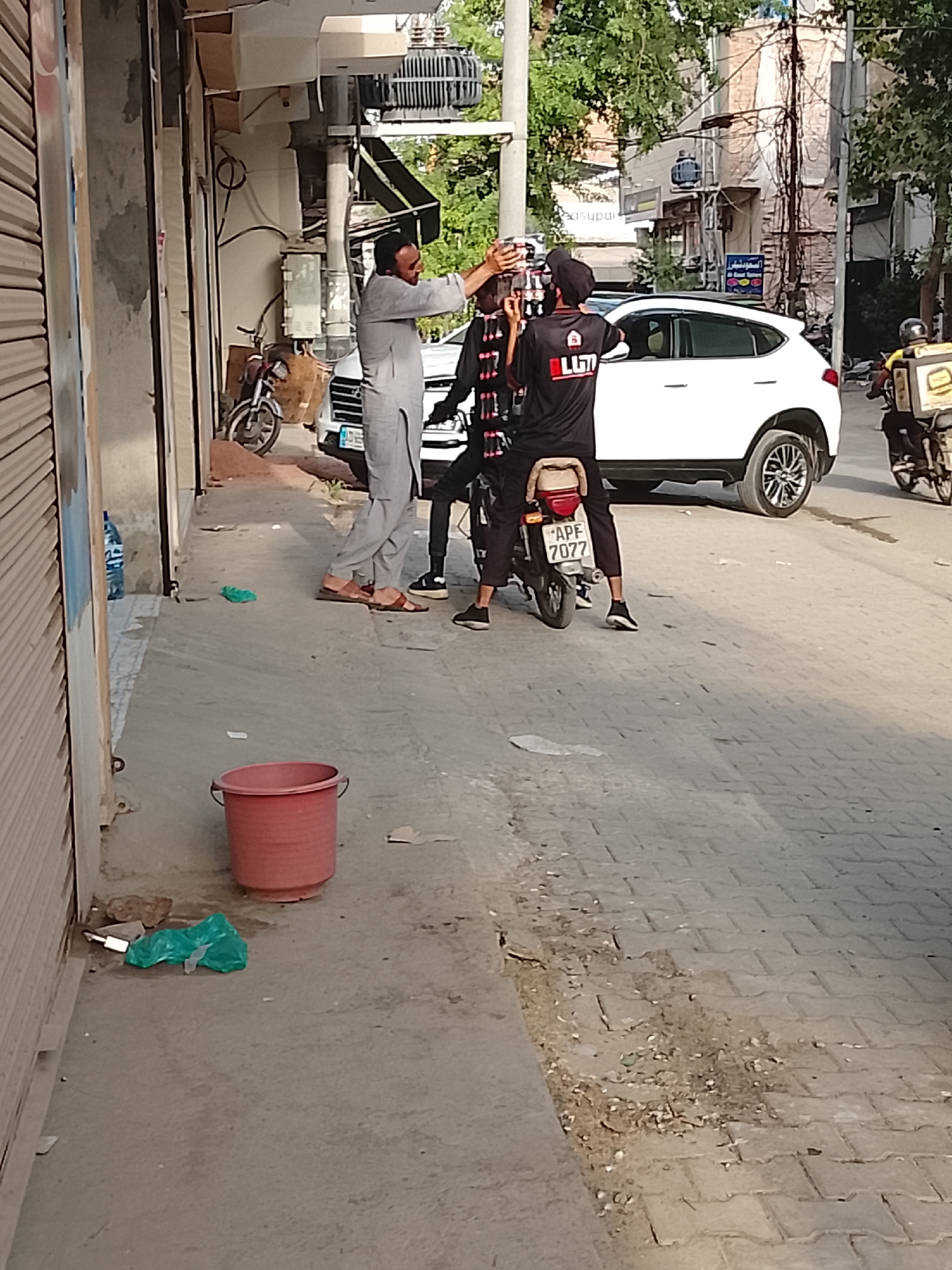 The image depicts a street scene in Gulgasht Colony, Pakistan. A man in traditional Pakistani attire is standing on the sidewalk facing a person sitting on a parked motorcycle. The man is reaching toward the seated person's head or hat. The seated person is wearing a black shirt with a logo and a cap. The motorcycle has a license plate reading "APE 7077". A white SUV is parked behind the motorcycle. In the background, another motorcycle, likely a delivery vehicle, is driving away. On the sidewalk, a red bucket and some discarded plastic are visible. The street appears to be a commercial area with buildings on either side. The lighting indicates it is daytime.