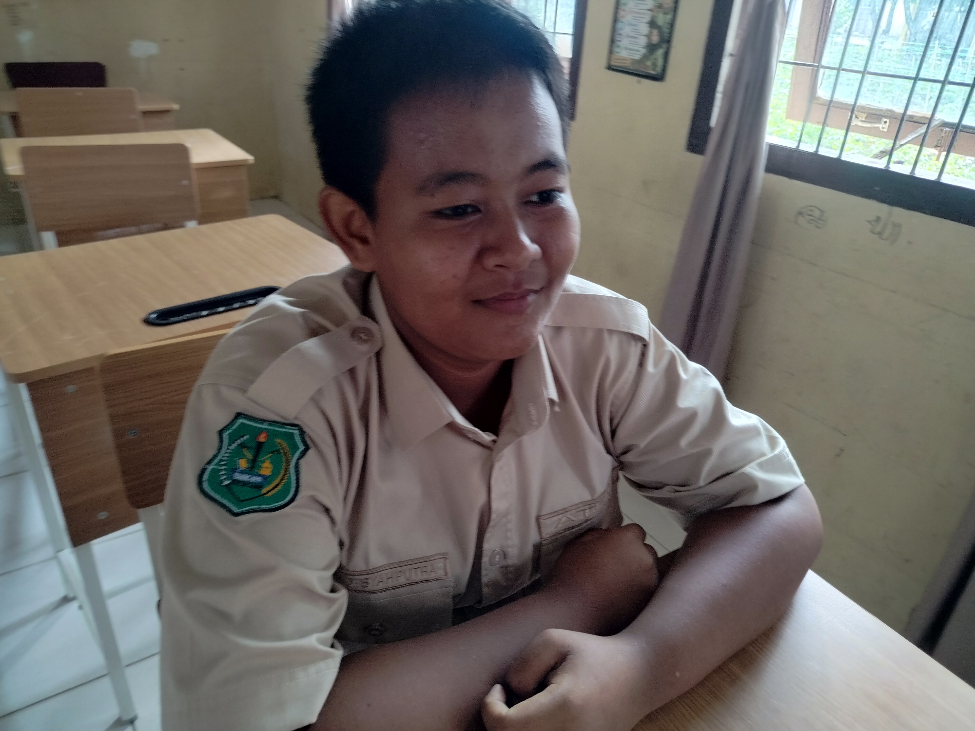 The image depicts a young person, likely a student, seated at a desk in what appears to be a classroom. The student is wearing a light-colored uniform shirt with an emblem on the left chest and a name tag on the right chest pocket. The background shows a typical classroom environment with multiple wooden desks and chairs, a window with a barred opening, and a light-colored wall. The overall scene suggests an indoor setting in a school. Based on the metadata associated with this image, the location is identified as Lawebekung, Indonesia.