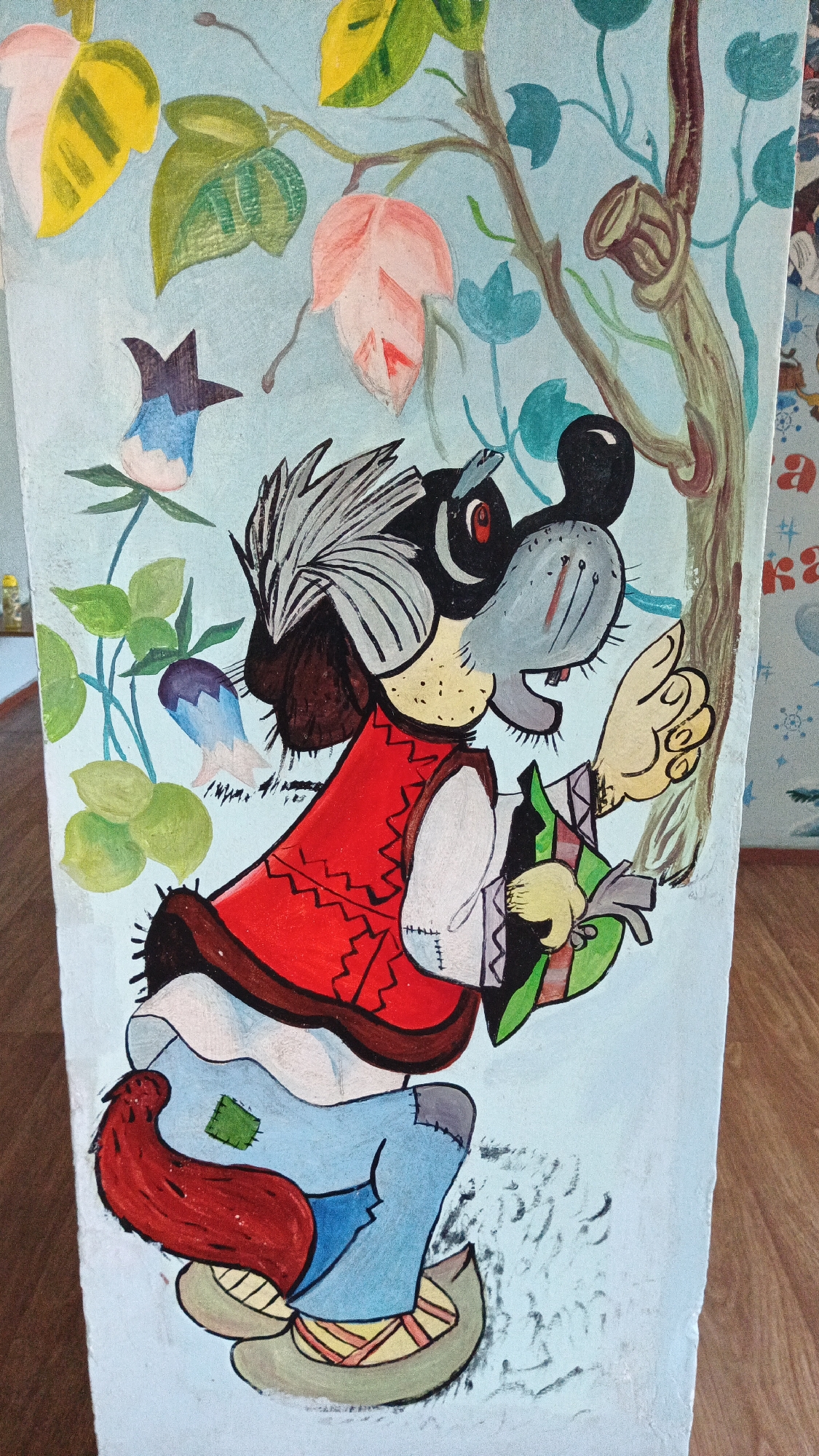 The image is a painting on a light blue background, depicting a cartoonish character that appears to be a dog wearing traditional clothing. The dog has a grey face with black ears, a black nose, and red eyes. It is wearing a red vest over a white shirt, blue pants with a green patch on the knee, and brown shoes. Its tail is red and bushy. The dog is holding a green object in its left hand. The background features a tree branch with green and pinkish-yellow leaves, and some blue flowers. The painting style is whimsical and childlike. The image is a photograph of this painting, with the painting appearing to be on a vertical surface, possibly a wall or a panel. Part of a wooden floor is visible at the bottom, and a patterned wallpaper is partially visible on the right side of the frame, with some Cyrillic letters visible on it. Based on the visible wallpaper pattern and Cyrillic lettering, this artwork is likely located in Kryvyi Rih, Ukraine.