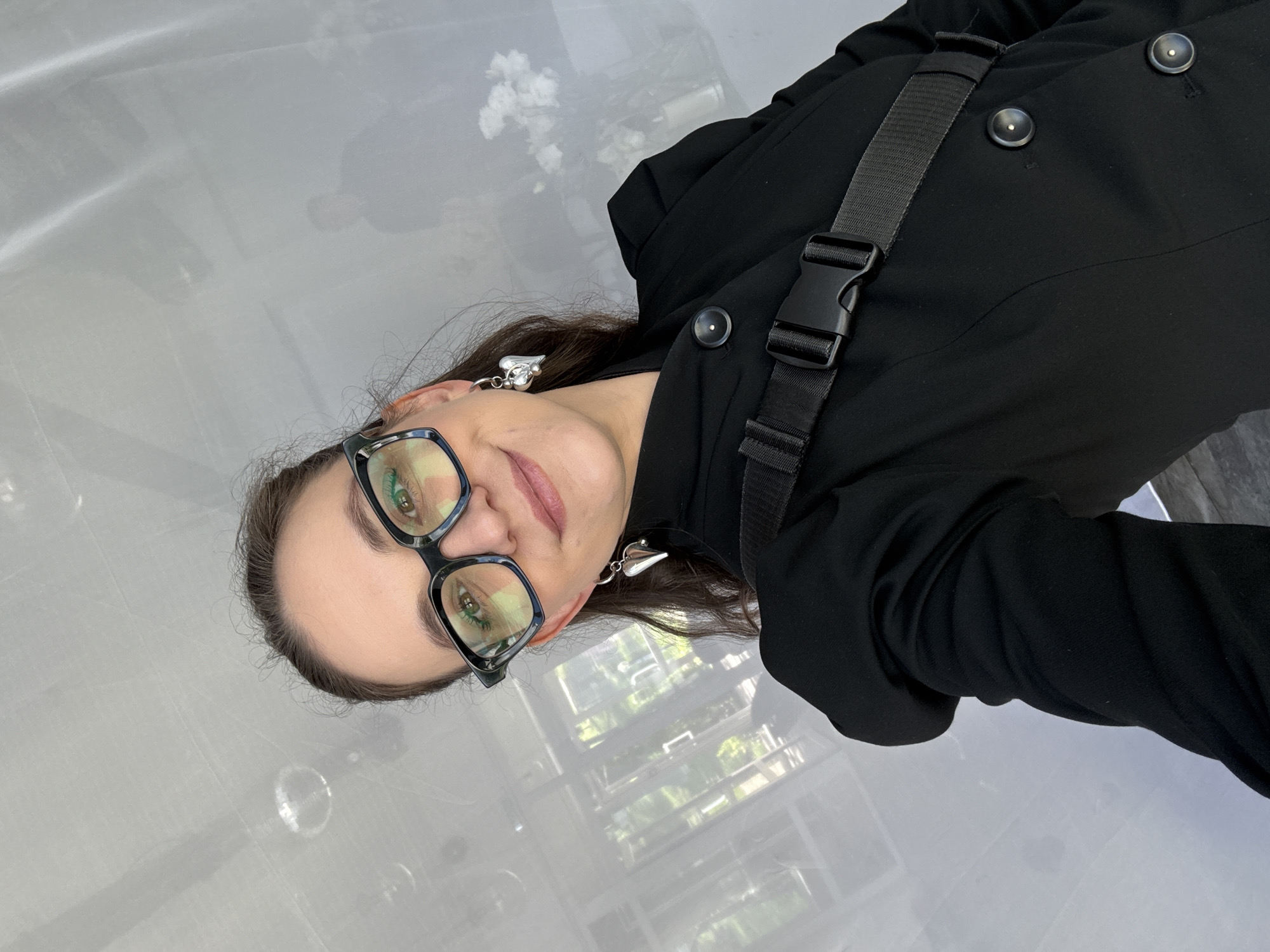The image is a selfie taken by a woman with dark hair and glasses, wearing a black jacket and heart-shaped earrings. She has a black strap with a buckle across her chest. The background appears to be an indoor space with a white, textured ceiling and a reflective surface that shows faint reflections of trees and a person. There are also blurred white flowers visible in the background. The woman is looking at the camera with a slight smile. The location is not identifiable as Votkinsk, Russia, or any other specific city.