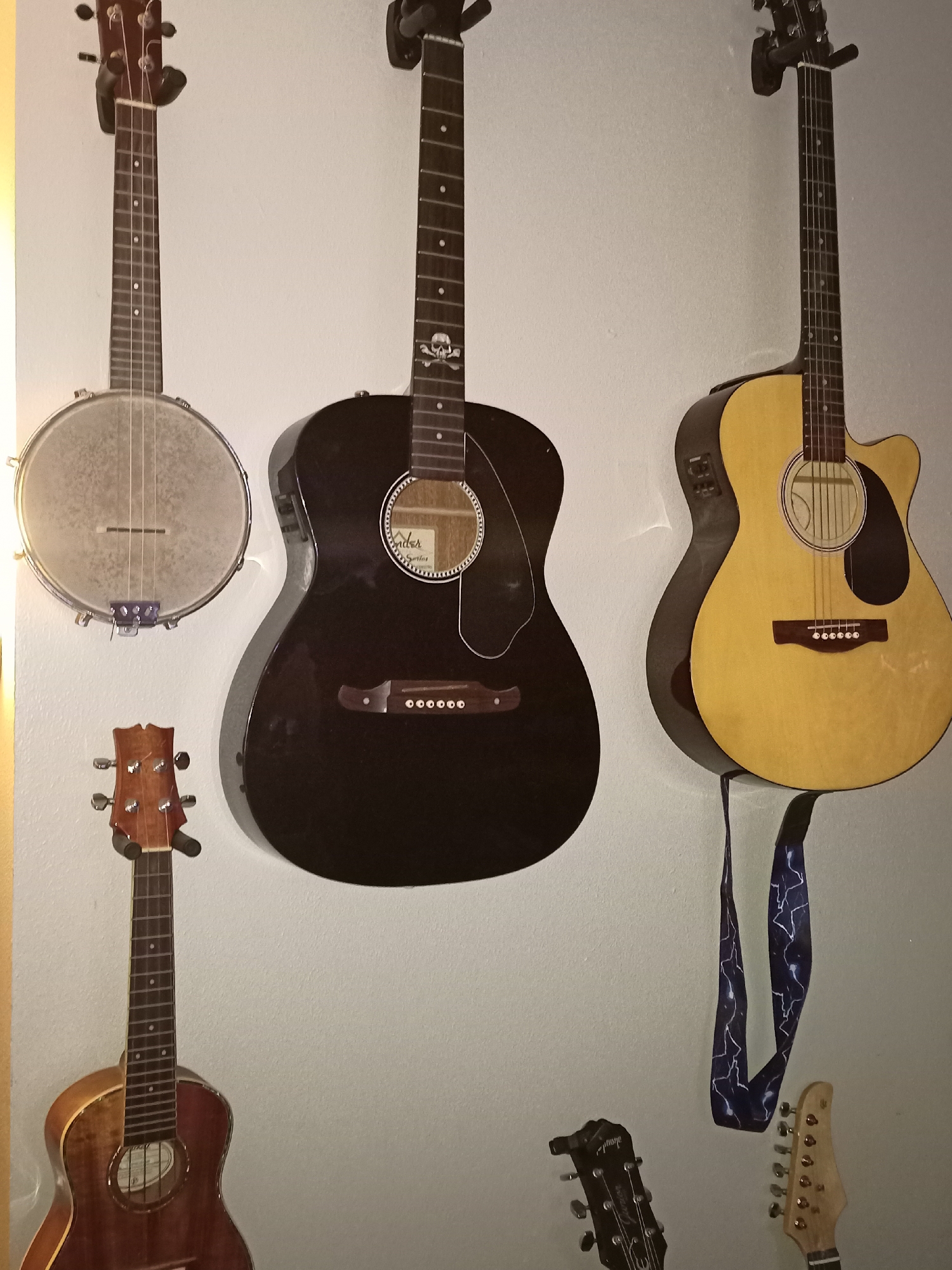 The image displays a collection of musical instruments, primarily guitars and a banjo, mounted on a light-colored wall. The instruments are arranged vertically and horizontally, creating a visually dynamic composition.
On the upper left, a banjo with a metallic resonator and a light-colored neck is suspended. To its right, a black acoustic guitar is hanging, featuring a white pickguard and a distinct skull inlay on the fretboard. Further to the right, a light yellow acoustic-electric guitar is mounted, distinguished by its cutaway body and a dark pickguard. Below the banjo and black guitar, a smaller, reddish-brown ukulele or small guitar is visible, with its headstock and tuning pegs on display. Towards the bottom right of the image, two guitar headstocks are visible: one appearing to be from an electric guitar and another, lighter-colored one. A guitar strap with a lightning bolt pattern dangles from the yellow guitar. The overall environment appears to be indoors, with soft lighting that casts subtle shadows on the wall. The image does not contain any discernible landmarks or contextual information that would place it in a specific city like Amarillo, United States of America.