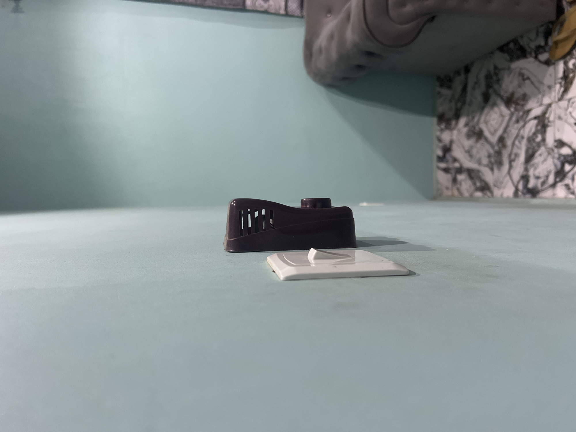The image shows a close-up of a wall in an indoor setting, likely a home or office. A white electrical switch plate is mounted on the light blue wall. Attached to the right of the switch plate is a dark brown electrical device, possibly a plug-in air freshener or a pest repeller, with ventilation slots and a small dial on its side. The wall extends to the right, revealing a corner where another wall, painted a similar shade of light blue, is visible. In the background, on the right side of the image, a portion of a tufted grey sofa is visible. The floor beneath the sofa is tiled with a black and white patterned design. A sliver of a yellow object, possibly a sandal, is visible at the bottom right edge of the frame. The lighting suggests it is daytime, with natural light entering the room. The scene does not provide any specific indicators to pinpoint its location within Jalingo, Nigeria, as it depicts common household items and decor.
