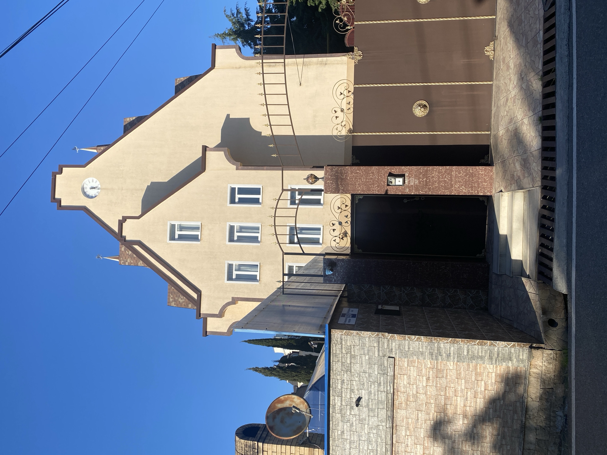 This media file presents an outdoor view of a multi-story building and its surrounding property in Sochi, Russia, captured on a clear, sunny day. The strong shadows cast suggest it is either morning or early afternoon.
The main subject is a light beige building with a distinctive stepped gable roofline, accented with dark brown trim. A circular white clock with Roman numerals, appearing to show approximately 9:15, is embedded in the upper part of the gable. Multiple rectangular windows with white frames are visible on the building's facade. An ornate metal structure, resembling a ladder or decorative element, extends upwards along the building's side from the gate area.
To the right of the building, a large, tall, brown metal gate with intricate gold-colored wrought iron scrollwork and circular medallions dominates a portion of the scene. Below the gate, a tiled pathway leads to an entrance area where a brown marble-textured panel houses an intercom or doorbell system. Faint, illegible text is visible on a small sign below a white box on the panel.
The ground in the foreground is covered with light-colored, patterned tiles, showing various shadows. A paved road with a drain runs along the right edge of the image. In the lower left, a large, somewhat rusty satellite dish and a brick structure, possibly an outdoor oven or chimney, are present. Green trees are visible in the background and over the gate. Power lines stretch across the clear blue sky in the upper left. No people are visible in the image.