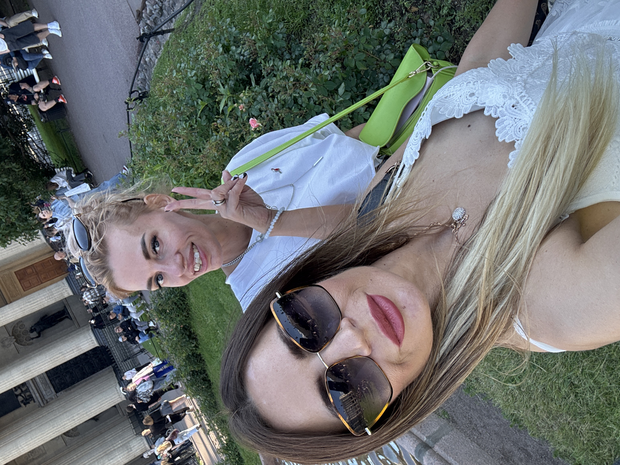 Two women are taking a selfie outdoors. The woman in the foreground on the right is wearing sunglasses and has long, blonde hair. She has on a white dress with lace detailing. The woman on the left has blonde hair and is wearing a white shirt and sunglasses. She is making a peace sign with her fingers. She has a green purse over her shoulder. The background shows a grassy area with some bushes and a building with columns, suggesting a public space or park. The lighting indicates it is daytime, likely late afternoon or early evening, given the soft light. The overall mood appears cheerful and relaxed, with both women smiling. The presence of columns and the style of the building in the background could be indicative of a historical or neoclassical architectural style, consistent with some areas in Saint Petersburg, Russia. No visible text is present in the image.