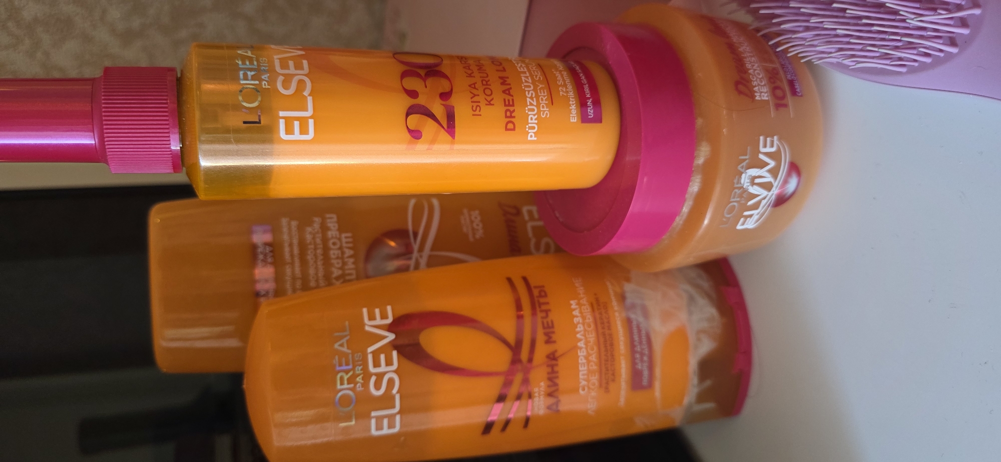 The image displays a collection of L'Oréal hair care products arranged on a light-colored surface. In the foreground, a round, orange L'Oréal Elvive hair mask with a pink lid is visible. Behind it and slightly to the left, an orange L'Oréal Elseve shampoo or conditioner bottle is positioned. To the right and slightly further back, a taller, orange L'Oréal Elseve spray bottle with a bright pink cap stands. Further in the background, another orange bottle, possibly a shampoo or conditioner, is partially obscured. A pink hairbrush with fine bristles is also present on the right side of the image, peeking into the frame. The lighting suggests an indoor setting, likely a bathroom or vanity area. There are no people or discernible city elements, making it impossible to determine its location within Sumgayit, Azerbaijan.
