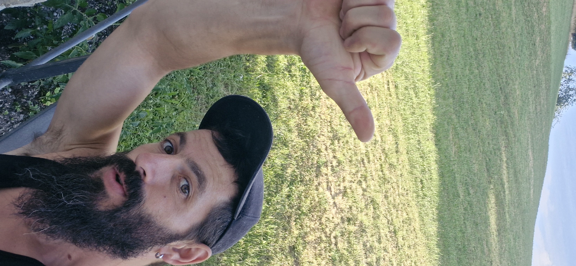 In an outdoor, grassy environment, a man with a beard and a black baseball cap is visible. He is looking at the camera with his mouth slightly open. His left arm is raised, and his thumb is extended upwards, creating a "thumbs up" gesture. The background consists of a sloping green lawn under a bright sky.