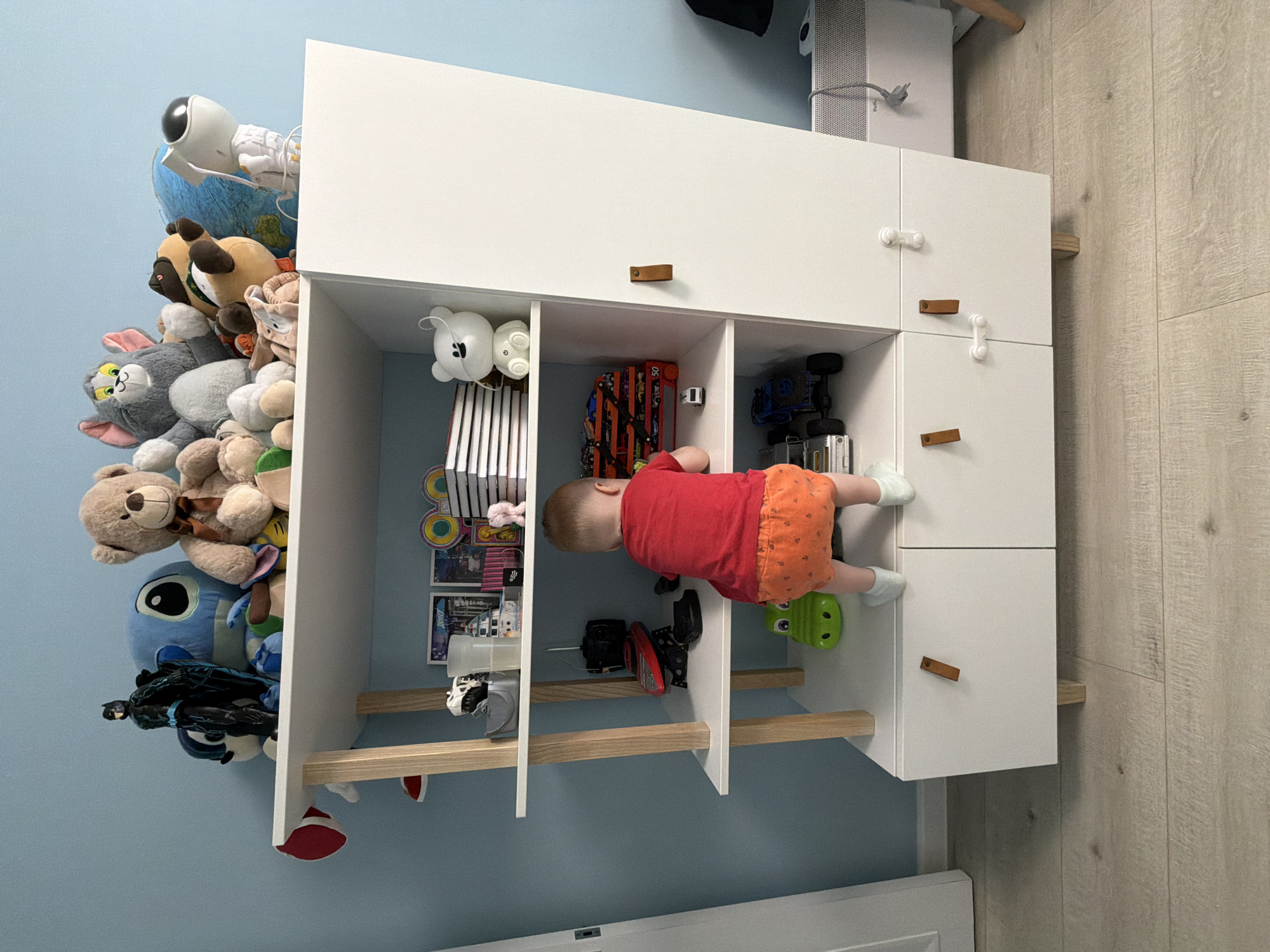 The image, taken in Votkinsk, Russia, captures a playful scene in what appears to be a child's room or play area. The central focus is a white modular storage unit with various compartments and cabinets, positioned against a light blue wall. To the right of the unit is a light wood laminate floor.
A young child, a toddler, is actively engaging with the storage unit. Dressed in a red short-sleeved top, orange patterned shorts, and white socks, the child is seen from behind, leaning into one of the open compartments on the lower left side of the unit. Their head and upper body are inside the shelf, suggesting they are reaching for or exploring items.
The storage unit itself is filled with toys. On top of the unit, to the left, there's a large pile of plush toys, including an astronaut figure standing on a globe, a grey Tom the cat plush, a brown teddy bear, and a blue Stitch plush toy. A Black Panther action figure is also visible among the plush toys lower down.
Inside the open shelves, various items are visible: white, vertically stacked books or boxes, several toy cars (including a blue truck and a red car), and a green alligator or dinosaur toy on the bottom shelf. Some closed cabinets have small wooden handles or white knob-like pulls. Wooden rungs or bars are integrated into the left side of the unit, possibly for support or as a climbing feature.
In the upper right corner, behind the storage unit, a white electronic device (possibly an air purifier or speaker) with a grey power cord is visible. The image is oriented sideways, with the wall appearing on the left and the floor on the right. No discernible text is visible on any of the objects. The scene conveys a sense of childhood exploration and play within a domestic setting.