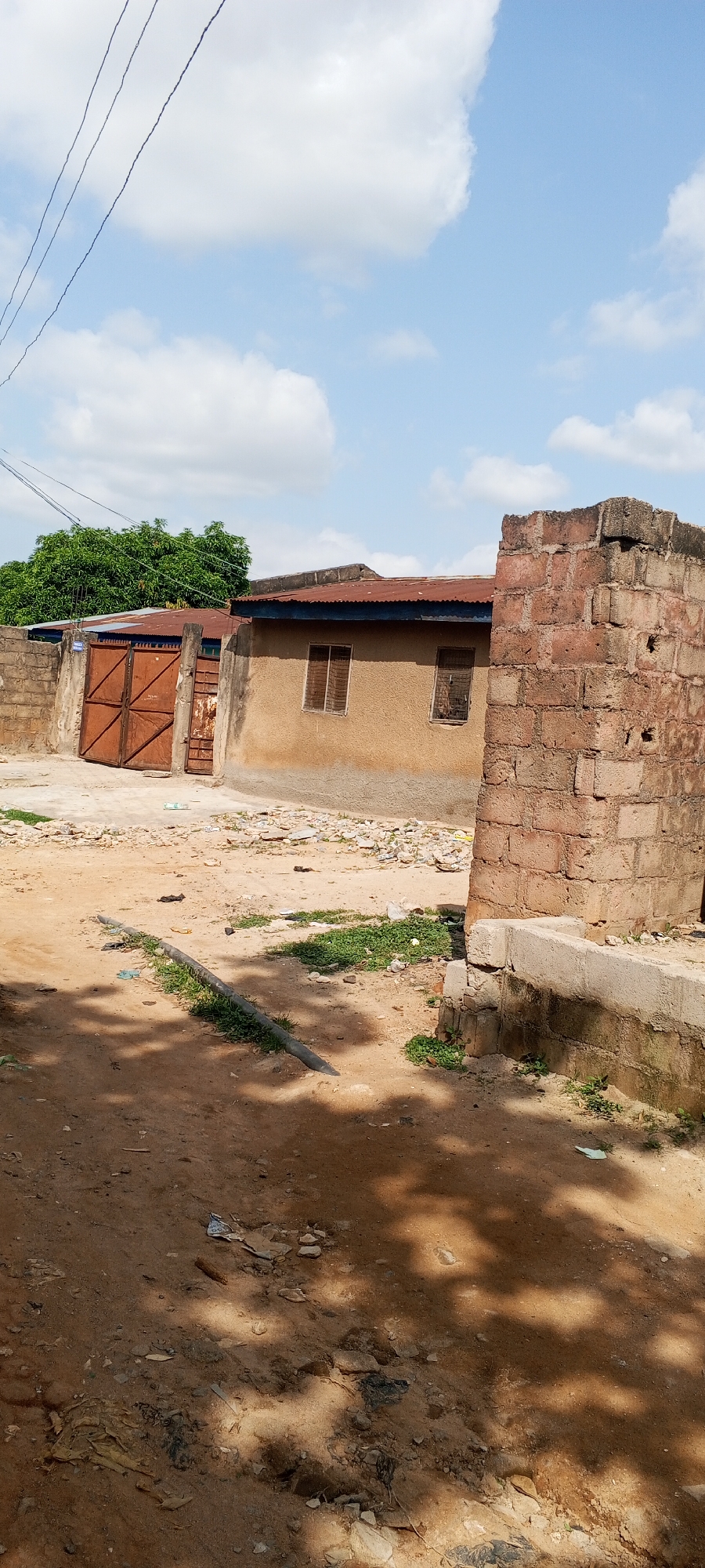 The image depicts an outdoor scene, likely in a residential area of Jalingo, Nigeria, characterized by modest construction and a somewhat unkempt environment. In the foreground, a dirt road with scattered debris and shadows cast by foliage is visible. A dark, cylindrical object, possibly a pipe, lies horizontally on the ground, partially overgrown with grass.
To the left, a rusty metal gate, with diamond-shaped patterns, is set within a brick wall. Behind the gate, a building with a corrugated iron roof and two windows with wooden shutters is partially visible, along with lush green trees.
Dominating the right side of the image is a tall, weathered brick wall, constructed from reddish-brown blocks, with a concrete cap. This wall appears to be a boundary marker. The house itself, situated between the gate and the brick wall, has a textured, light brown stucco exterior and a corrugated iron roof. It features two rectangular windows, also with wooden shutters. The ground around the structures is a mix of dirt and scattered refuse, suggesting a lack of recent maintenance. The sky above is blue with scattered white clouds, and electrical wires are strung across the upper portion of the frame.