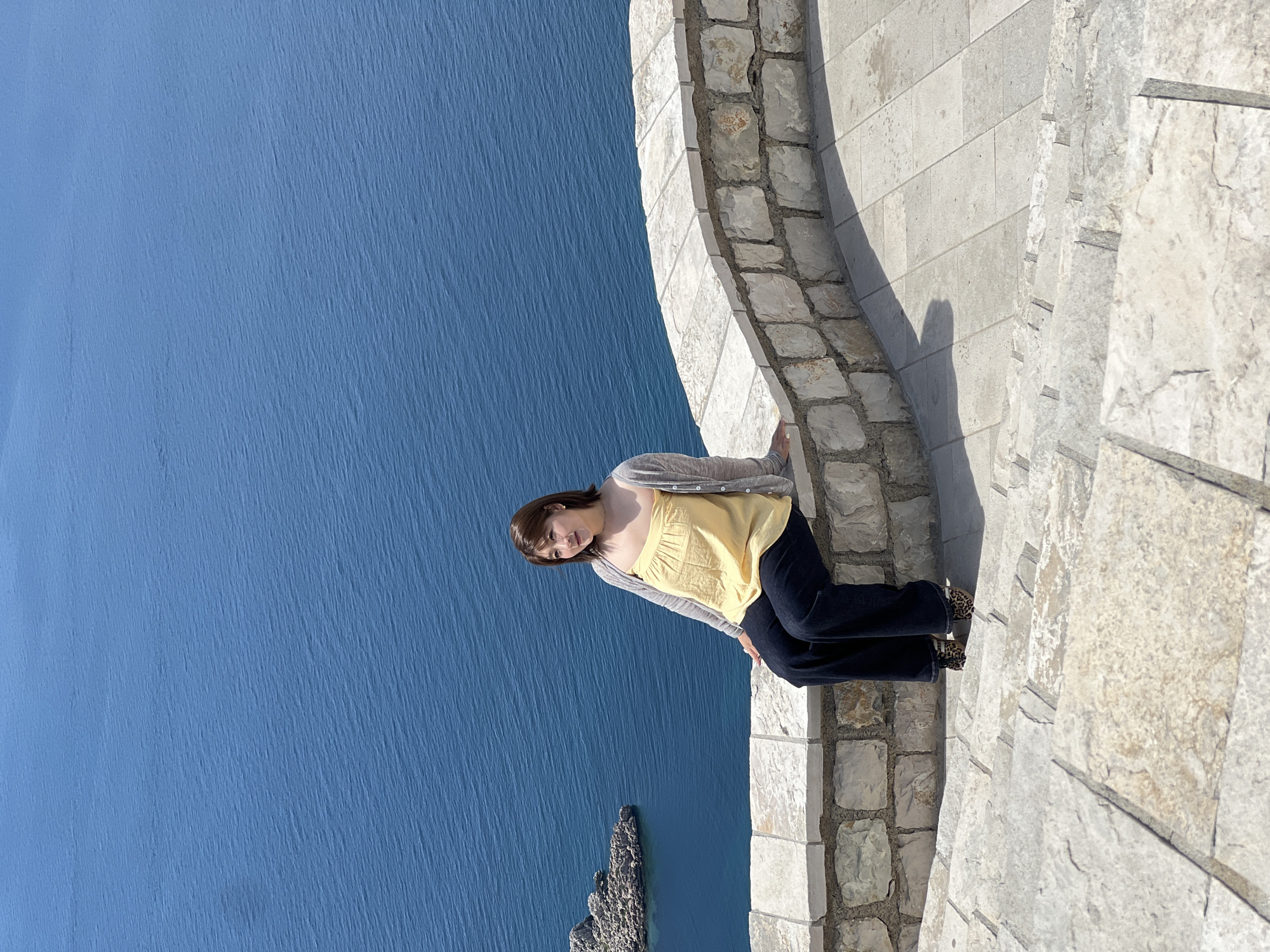 A woman stands on a stone path overlooking the Adriatic Sea at Sveti Stefan, Montenegro. The woman has shoulder-length brown hair, wears a yellow tank top, a light gray cardigan, and dark pants. She is standing on a path constructed of irregular light-colored stone blocks. The path is edged with a low stone wall. To the left of the path, the blue expanse of the Adriatic Sea stretches out, punctuated by a rocky islet in the foreground. The sky is not visible. The image is rotated approximately 90 degrees counterclockwise.