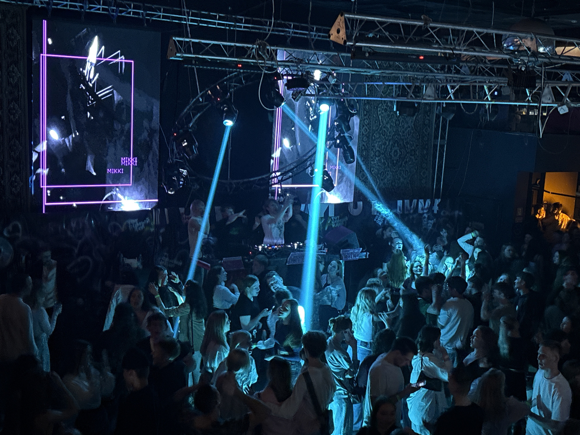 The image shows a crowded event in Izhevsk, Russia. A stage is set up with a large screen displaying graphics and the name "MIKKI" with blue spotlights shining on the audience. People are densely packed, seemingly enjoying the event, with some holding up phones and raising their hands. The environment appears to be a nightclub or concert venue, with dark walls and lighting equipment visible above the crowd.