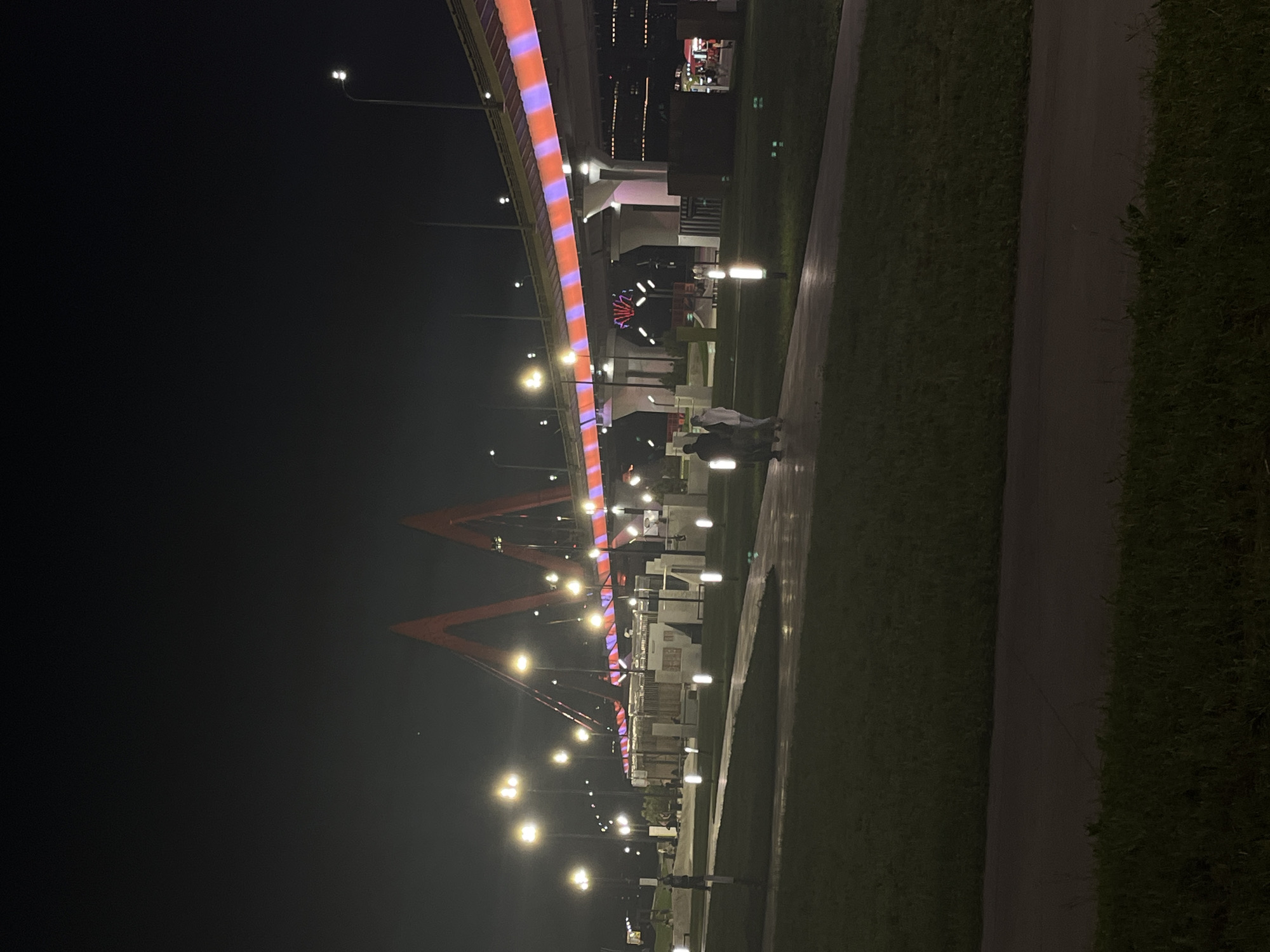 The image depicts a night scene in Kazan, Russia, featuring a bridge with orange and purple striped lighting spanning across a dark sky. Under the bridge, buildings and lampposts line a walkway. A few people are visible on the path. The sides of the walkway are flanked by grass.
