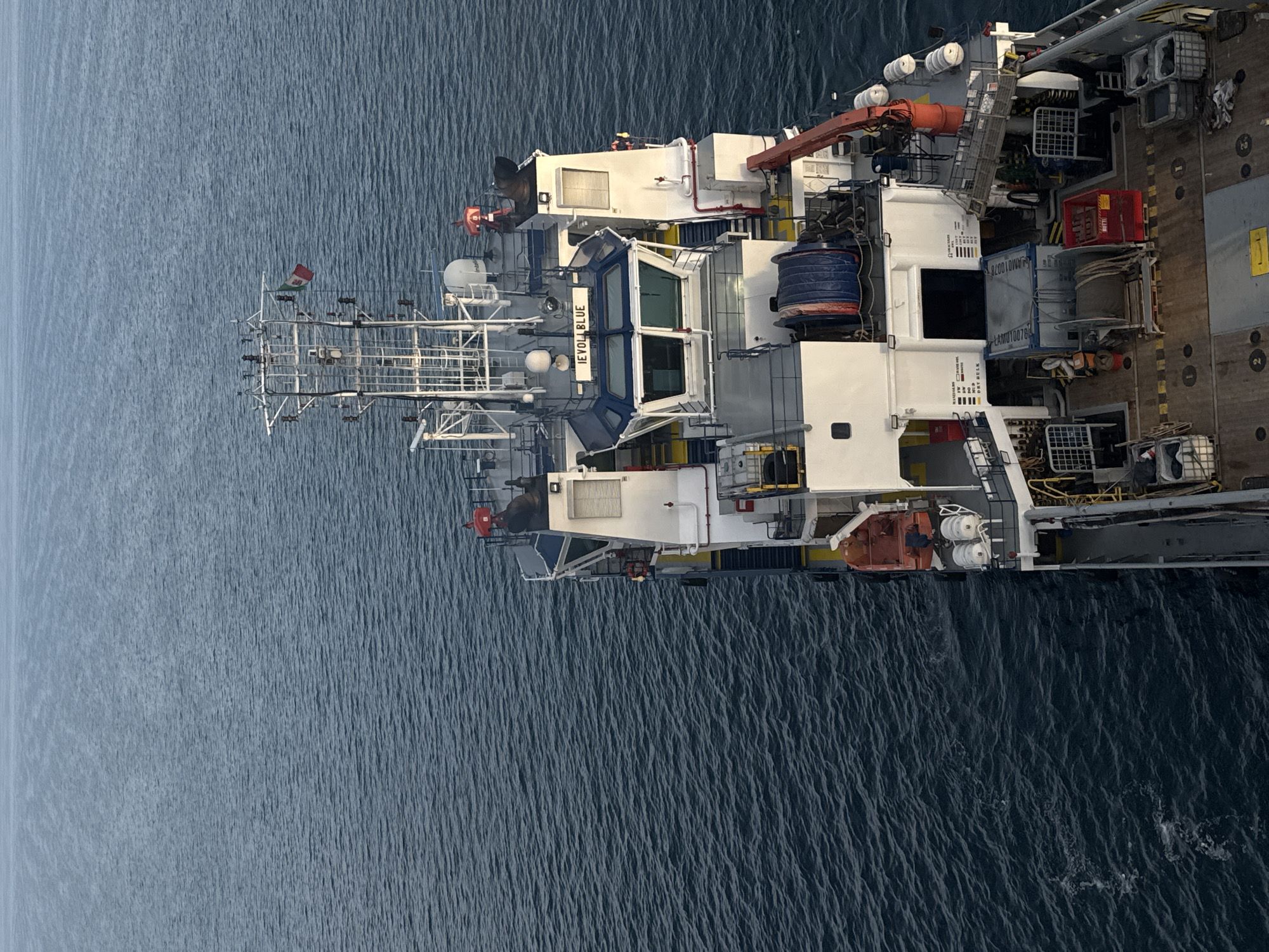 This is an aerial, high-angle photograph of a vessel on the water. The water is dark blue and appears to be choppy with small waves. The vessel is primarily white with some blue and red accents. It is oriented vertically in the frame, with the bow pointing towards the top left.
The vessel has multiple levels and a complex superstructure, including a bridge with large windows labeled with the text "NEYOLLA BLUE" in white lettering on a blue background. There are various pieces of equipment visible on the deck, including cranes, reels of rope or cable, and what appear to be storage containers or baskets. A flag, predominantly red and white with a green stripe, is flying from a mast on the forward section of the vessel. The time of day appears to be daytime, with no strong shadows to indicate early morning or late evening. The weather is likely clear, as there are no visible clouds. The setting is an open body of water, likely the ocean, given the size of the vessel. There are no people visible in the photograph. The overall impression is one of an industrial or working vessel at sea.