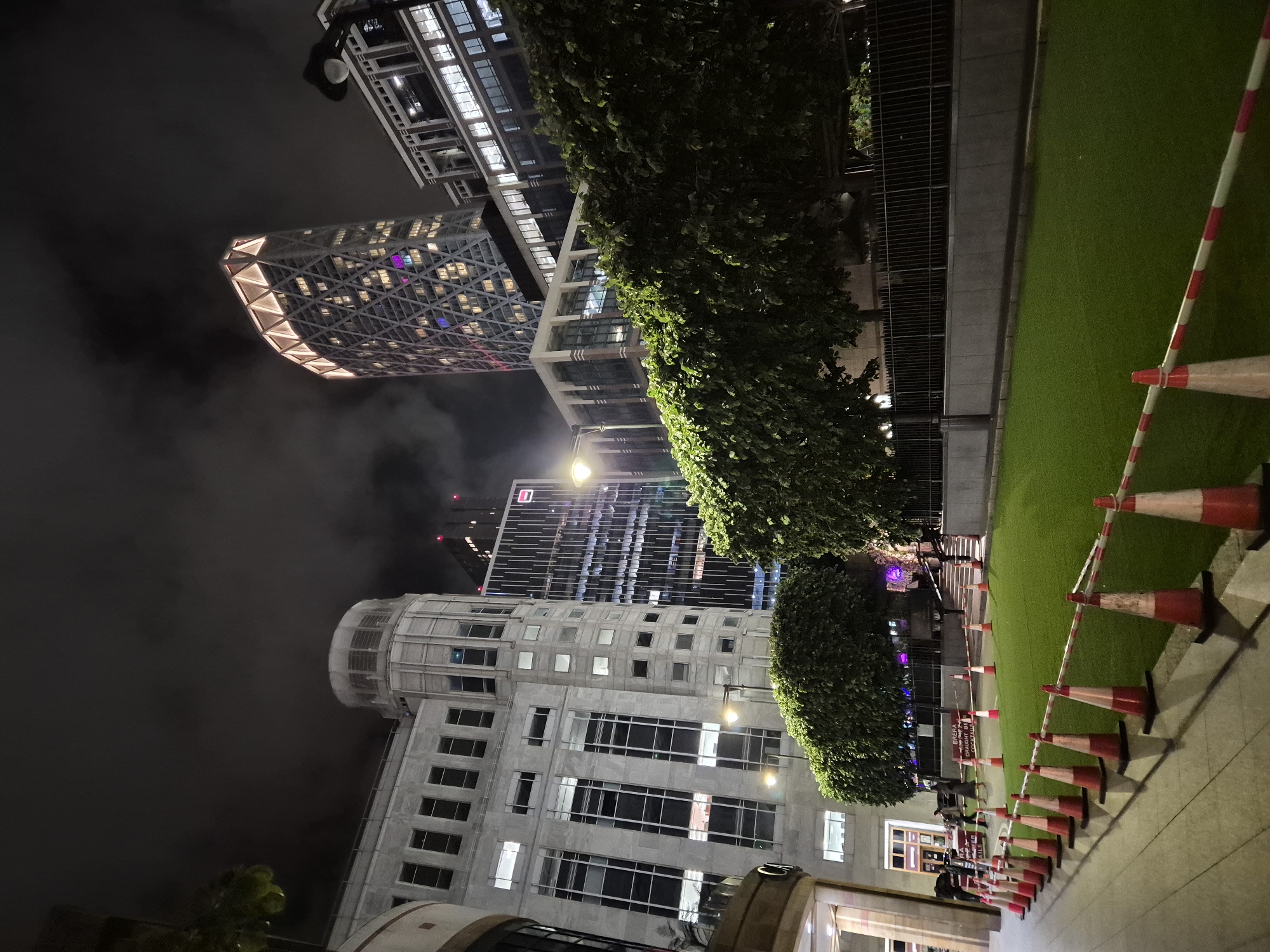 A nighttime, low-angle view in the City of London shows a line of traffic cones bordering a patch of artificial turf along a walkway. The image includes multiple tall buildings illuminated against a dark sky. Several skyscrapers are visible, including one with a distinctive angular, lattice-like exterior illuminated with warm lighting at the top left. Another is a modern skyscraper with a dark glass facade. Other buildings feature a more traditional, white-stone architecture. Trees line the walkway. Streetlights and building lights cast a glow on the scene. No people are clearly visible in the image, although figures can be discerned in the background.