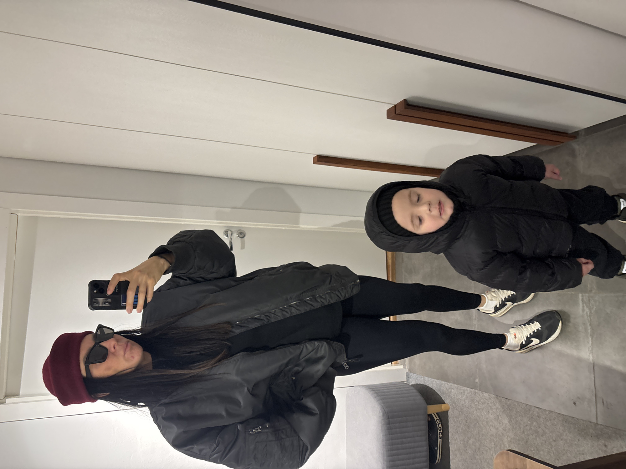 The image depicts a woman and a child, presumably in an indoor setting. The woman is holding a phone, likely taking a selfie, and is wearing a maroon beret, sunglasses, a black bomber jacket, and black leggings, with white and black sneakers. The child is lying on the floor in a black puffer jacket and black pants, with matching sneakers. The background suggests a modern interior with a white paneled wall, a gray floor, and minimalist wooden accents. It is not possible to tell from this photo that this is in Chaikovsky, Russia.