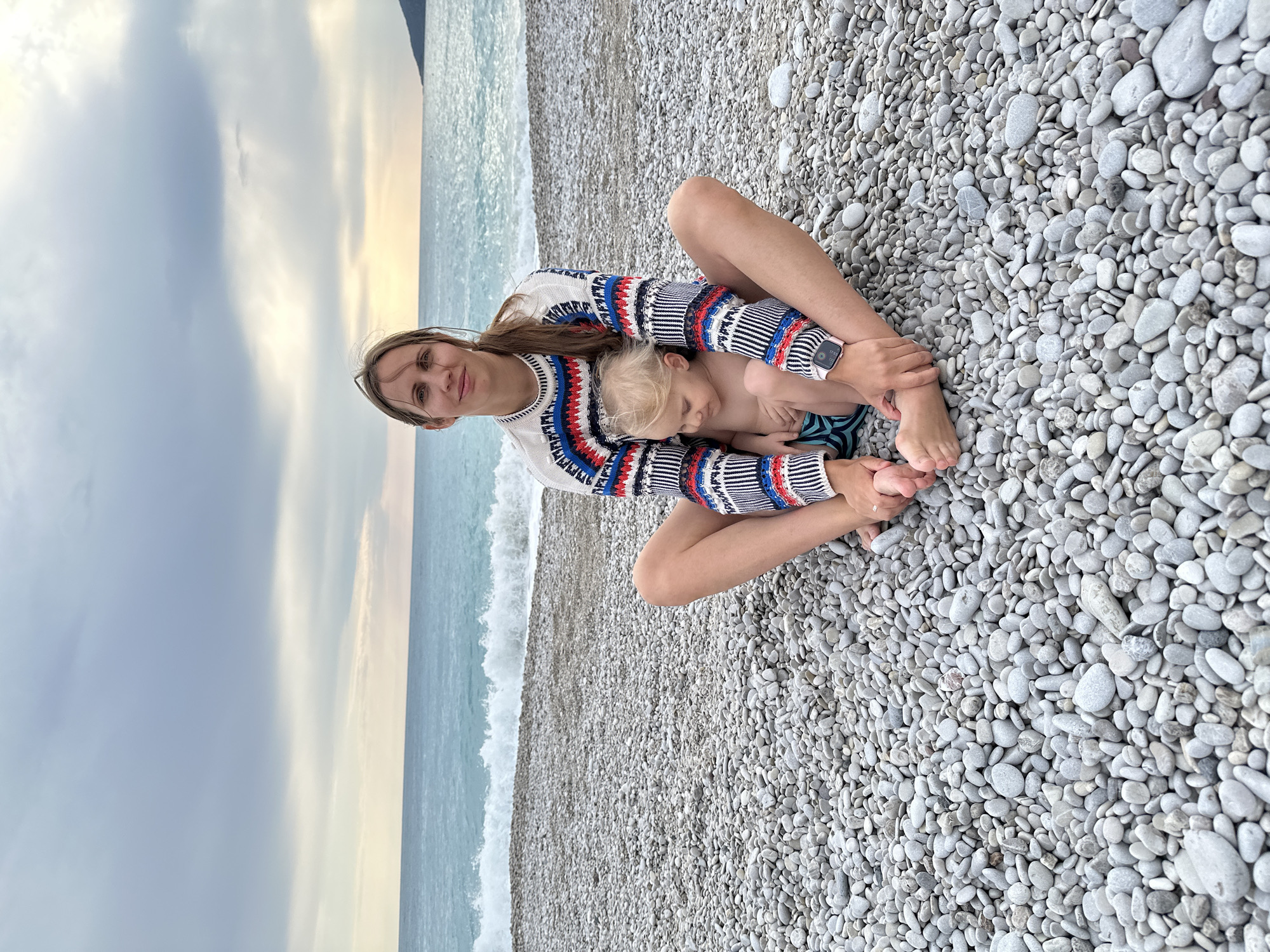 A woman in a colorful sweater sits cross-legged on a pebble beach with a young child resting against her. They are located in Fethiye, Turkey. The child is wearing blue and black striped swimwear. The sea and sky are visible in the background.